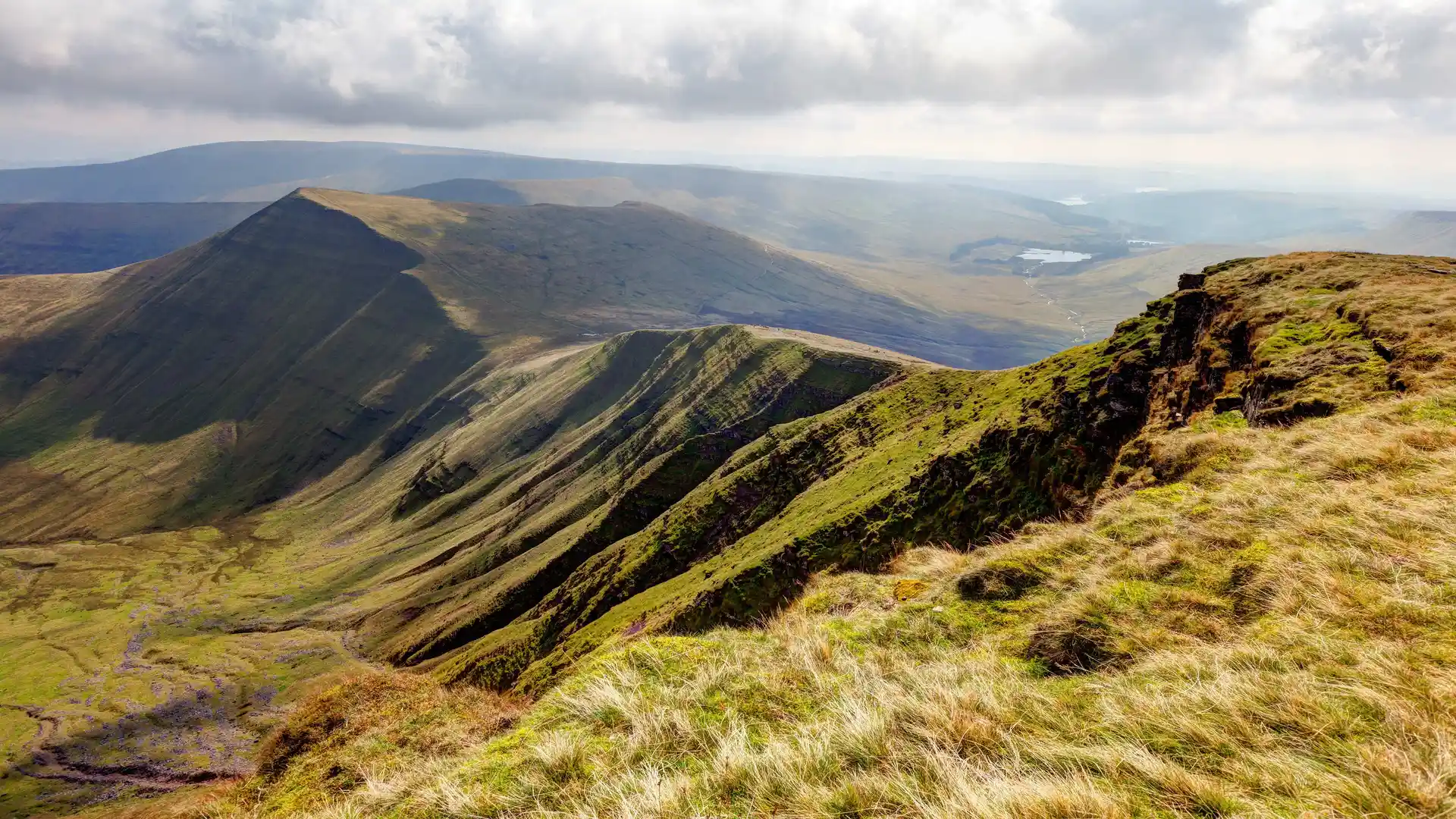 View from a high ridgeline in Bannau Brycheiniog (Brecon Beacons), South Wales, with steep grassy slopes, deep valleys, and distant reservoirs under a cloudy sky.