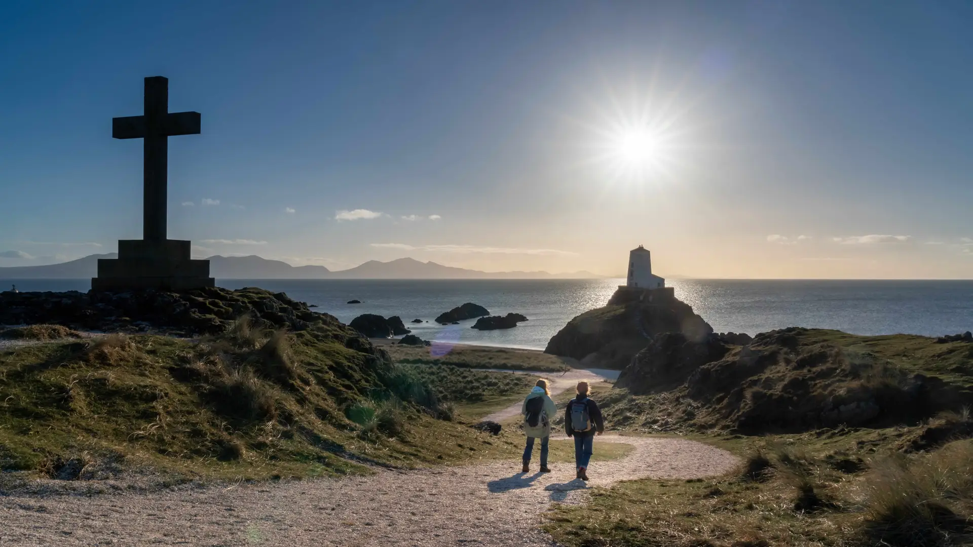 Walkers approaching Tŵr Mawr lighthouse on Llanddwyn Island, Isle of Anglesey, with Celtic cross, coastal views, and golden evening light.