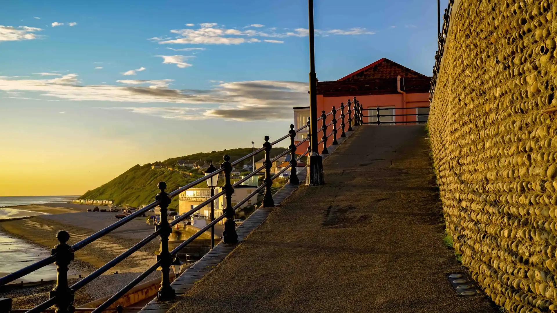 Seafront promenade in Cromer at sunset, with views over the beach, iron railings, and a flint wall leading towards the cliffs and houses beyond.