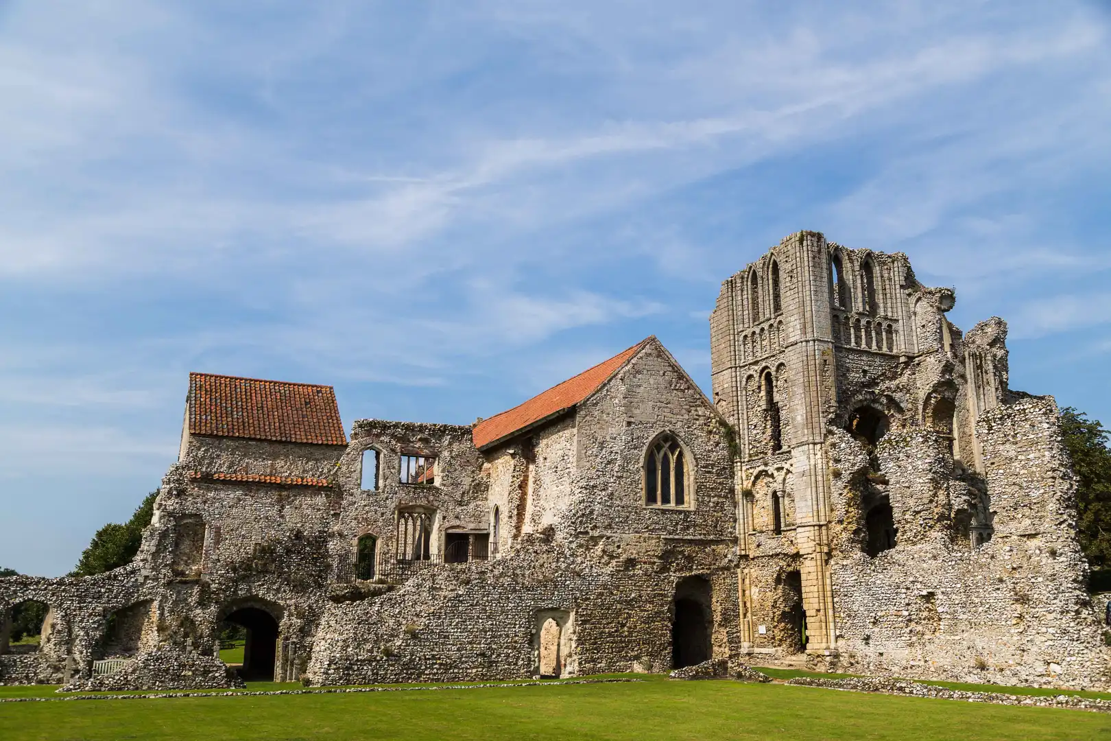 Ruins of Castle Acre Priory in Norfolk, with flint stone walls, arched windows, and a tall tower against a blue sky.