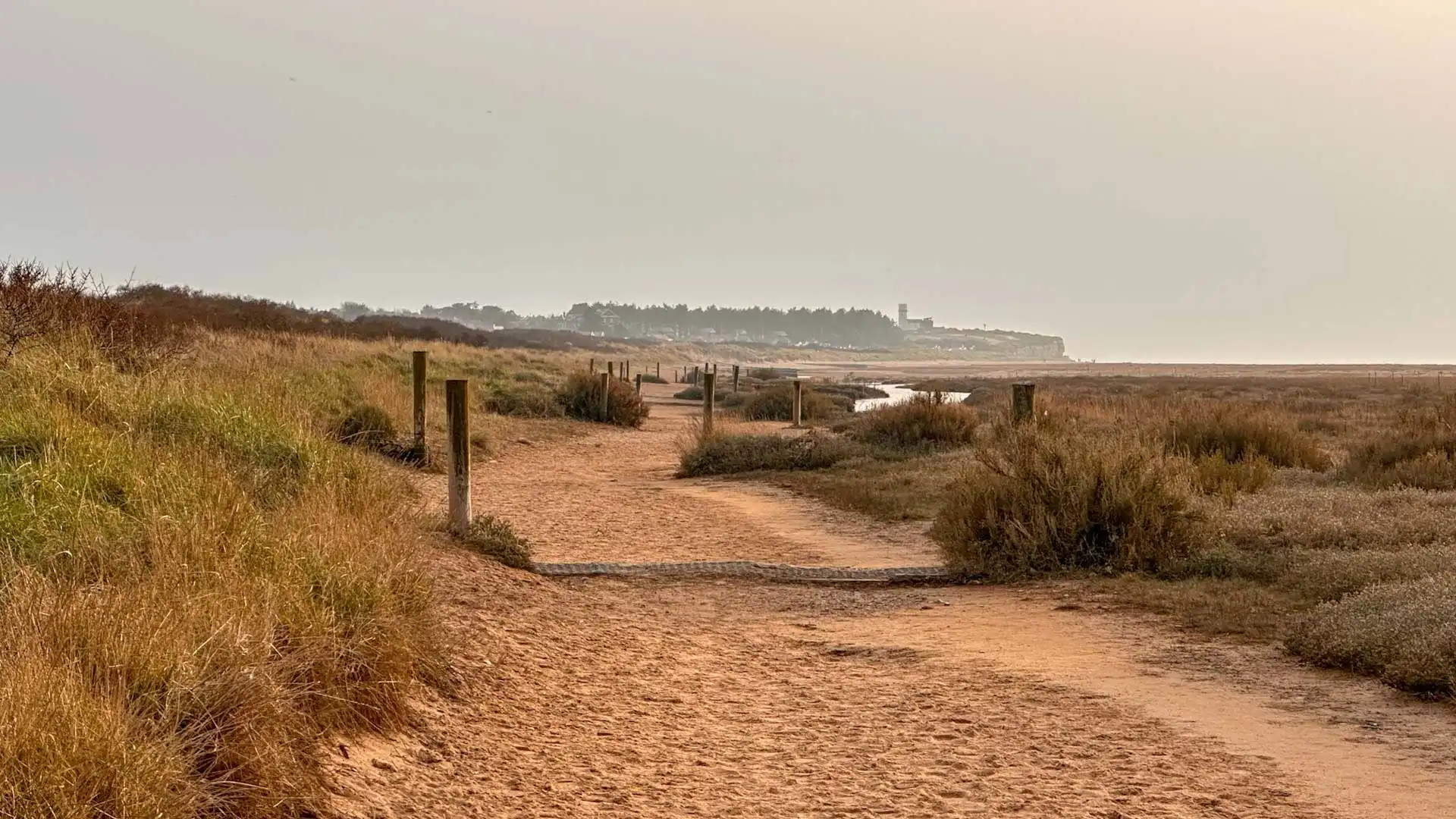 Sandy coastal footpath leading through dunes and saltmarsh at Holme-next-the-Sea, with distant cliffs and a soft, hazy sky.