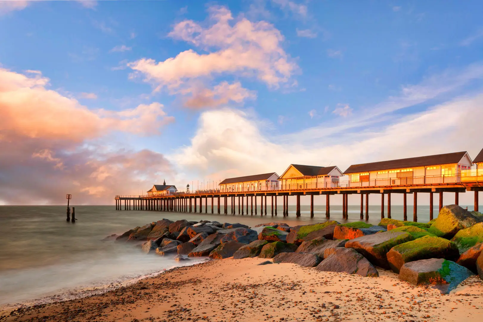 Southwold Pier at sunset, with warm light reflecting off its white wooden buildings. The pier stretches out into calm sea waters, framed by a sandy beach and moss-covered rocks in the foreground, under a sky of pastel clouds.