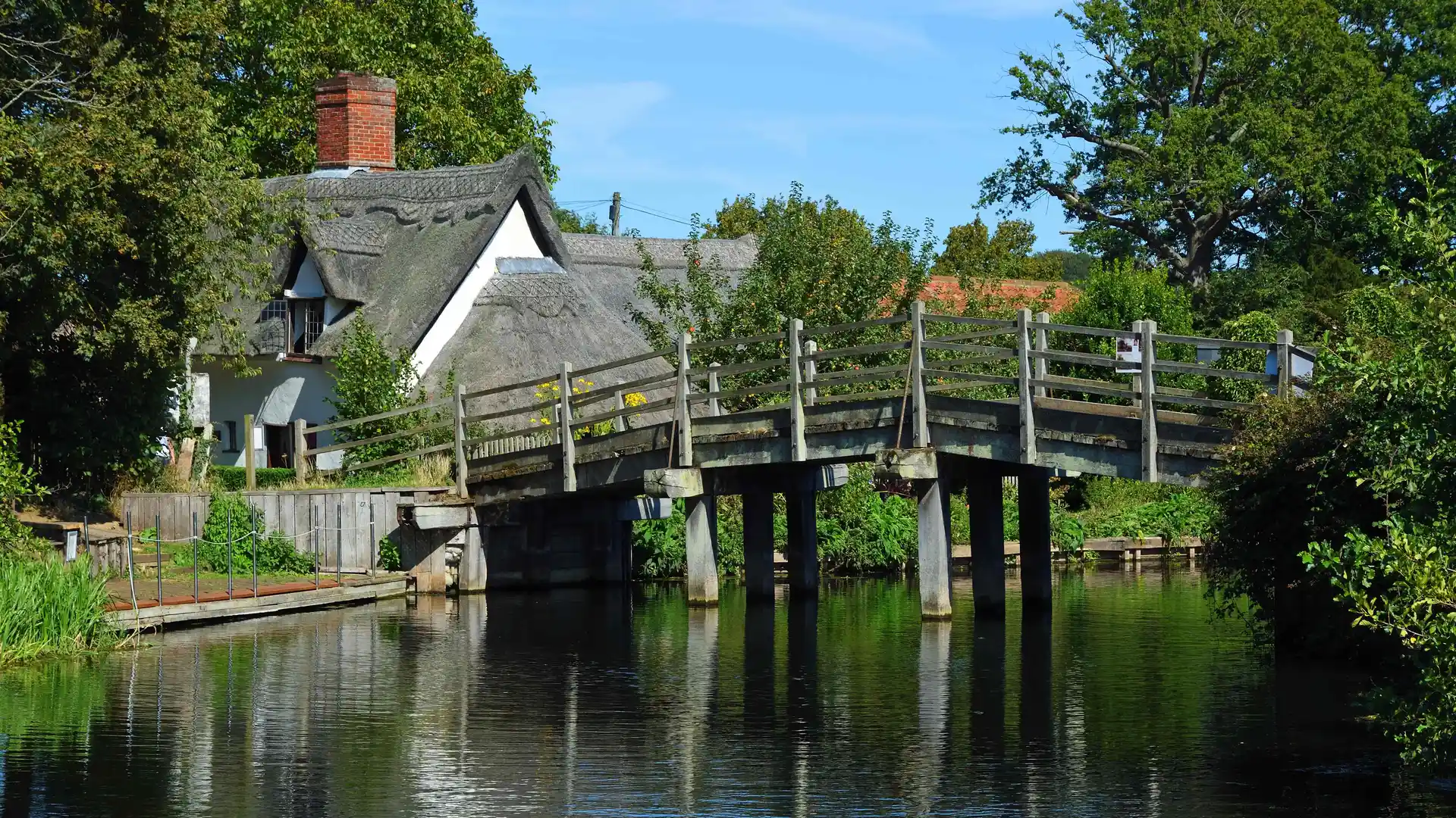 Wooden footbridge crossing the River Stour at Flatford, Suffolk, with a white thatched cottage and trees in the background on a sunny day.