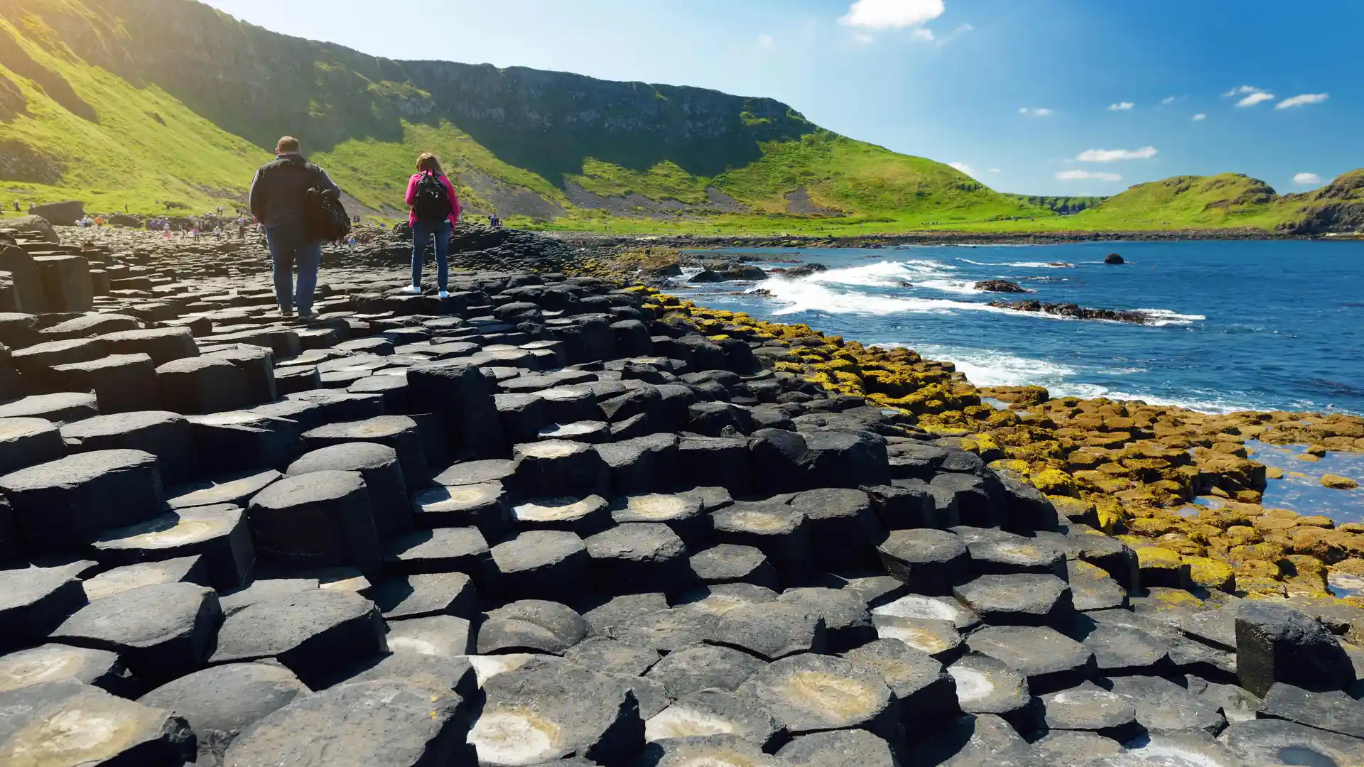 Two people walk across the hexagonal basalt columns of the Giant’s Causeway on Northern Ireland’s Causeway Coast, with green cliffs and a bright blue sea under a sunny sky.