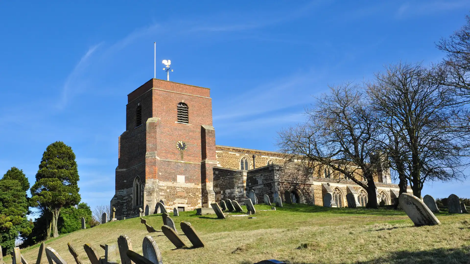 Historic church on a grassy hill with leaning gravestones in the foreground, Shillington, Bedfordshire, under a clear blue sky.
