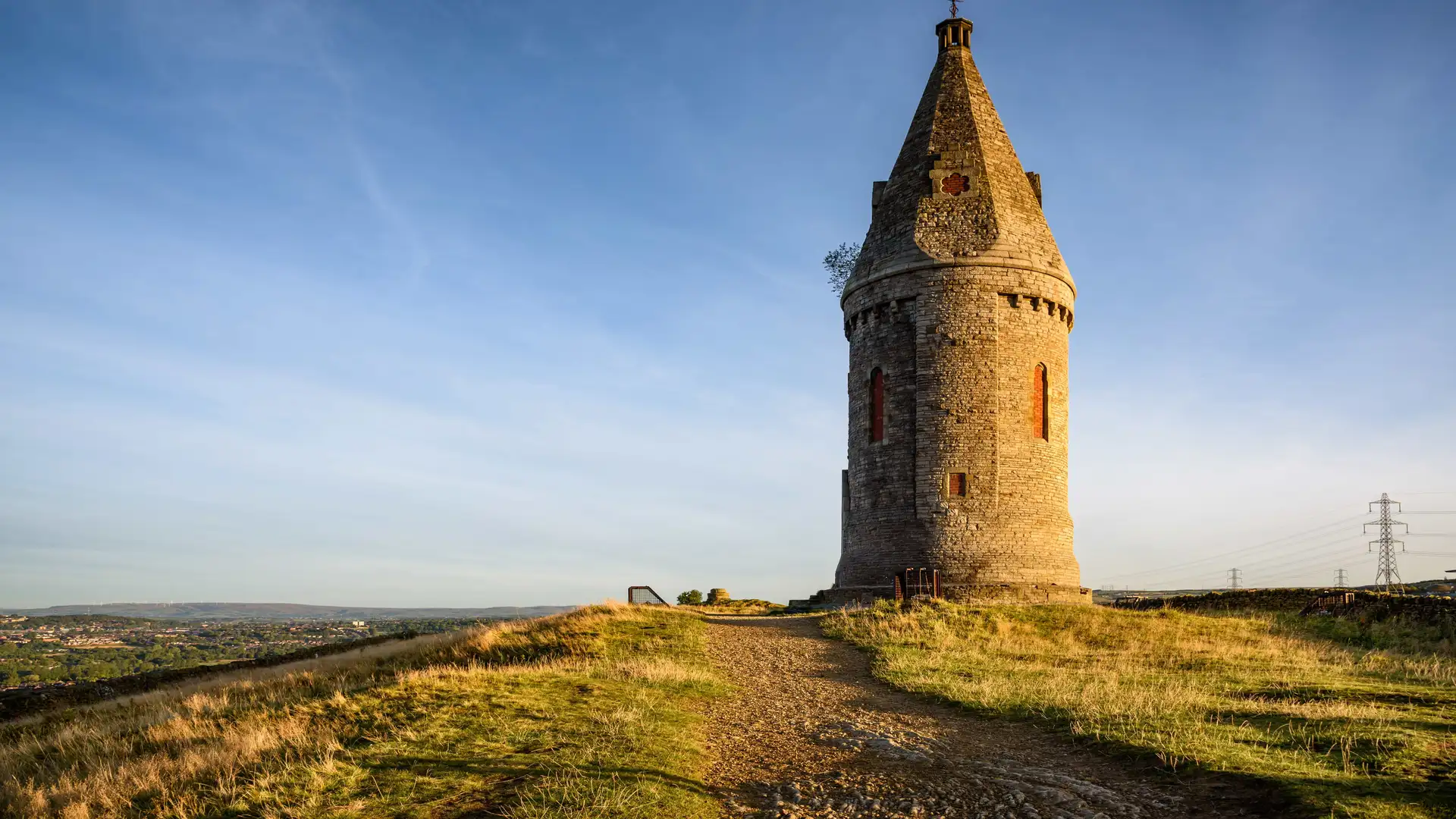 Hartshead Pike stone tower with walking trail in foreground, overlooking the Greater Manchester skyline on a clear day.