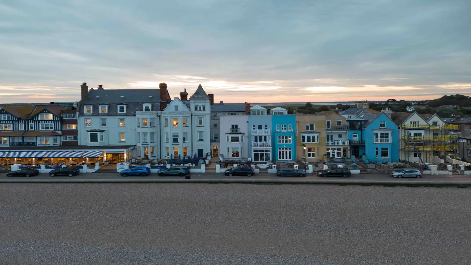 Seafront houses in Aldeburgh, Suffolk, at dusk — a row of colourful and historic buildings facing the shingle beach, with cars parked along the promenade and a soft sunset in the background.