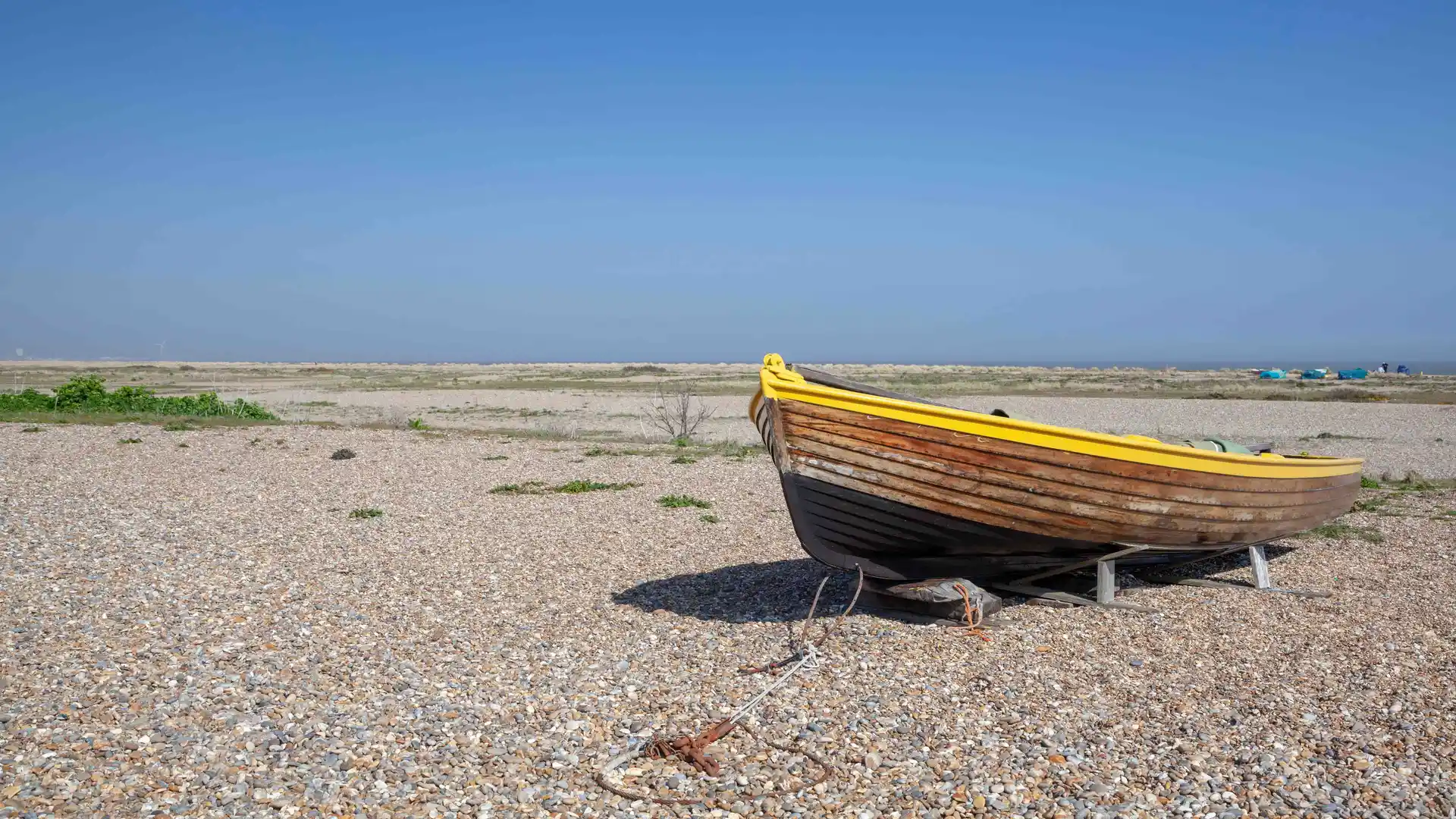 Wooden fishing boat with a yellow trim resting on a wide shingle beach at Kessingland, Suffolk, under a clear blue sky.