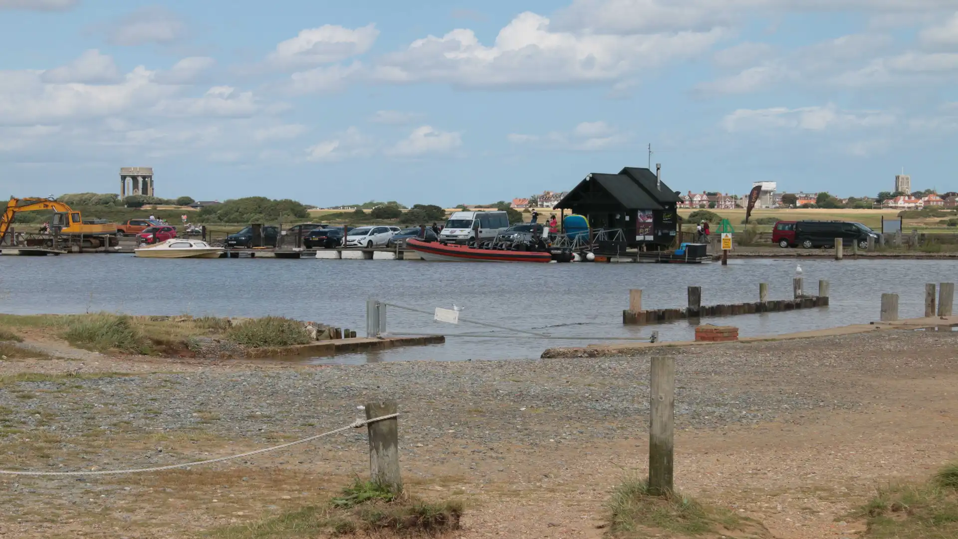 View across the River Blyth from Walberswick, showing the hand-pulled ferry, docked boats, and Southwold in the distance under a bright sky.