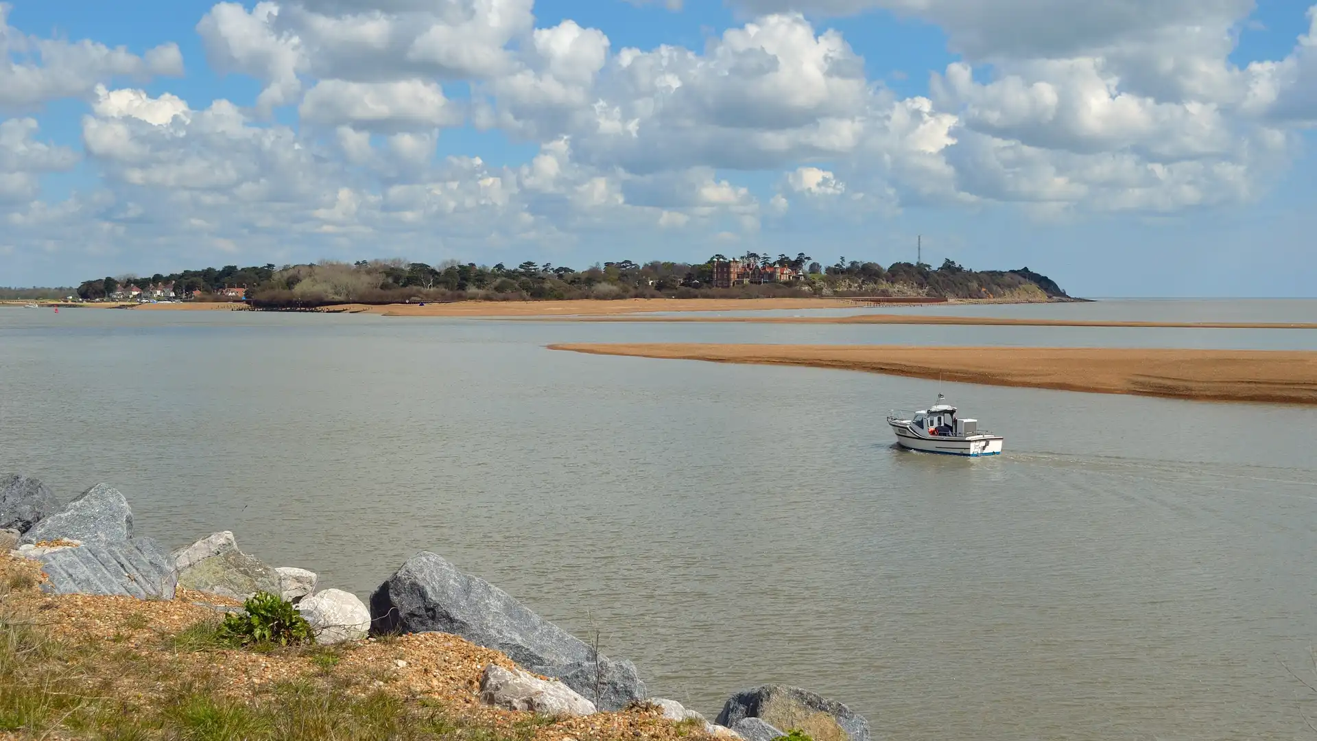 View across the River Deben towards Bawdsey cliffs and buildings, with a small boat on the water and wide coastal skies.