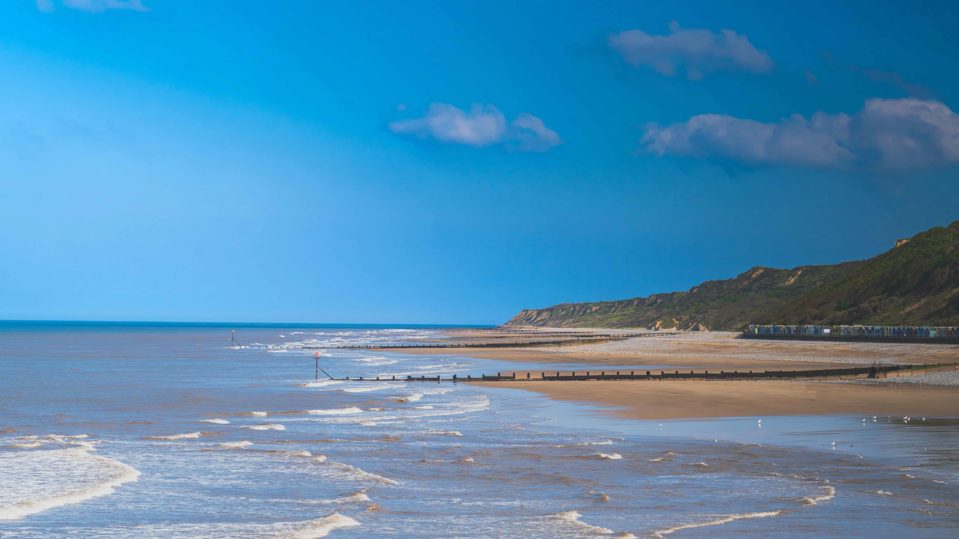 View along the beach at Overstrand, Norfolk, with wooden groynes, waves rolling in, colorful beach huts, and coastal cliffs under a bright blue sky.