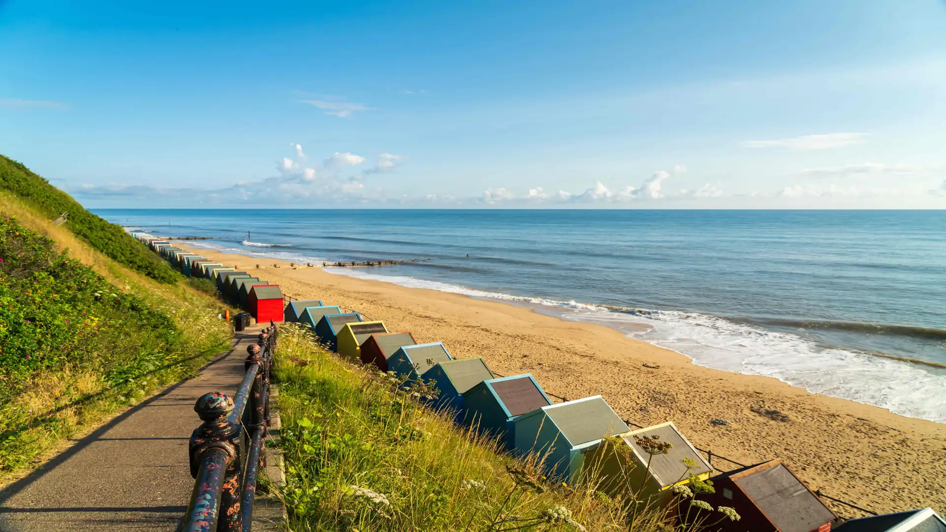 View down a path lined with colorful beach huts leading to the sandy beach at Mundesley, Norfolk, with calm blue sea and clear skies.