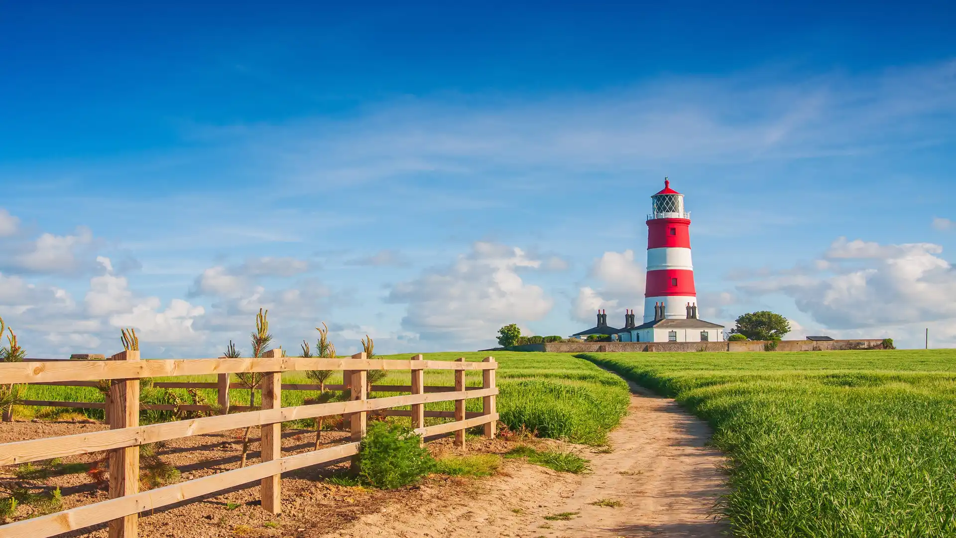 View of Happisburgh Lighthouse with red and white stripes, set against a blue sky and surrounded by green fields and a sandy footpath.