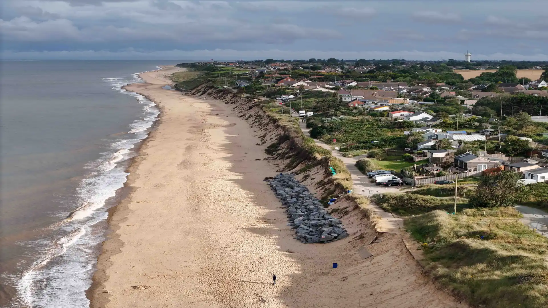 Aerial view of Hemsby Beach with golden sand, grassy cliffs, and seaside cottages stretching along the Norfolk coast under a cloudy sky.