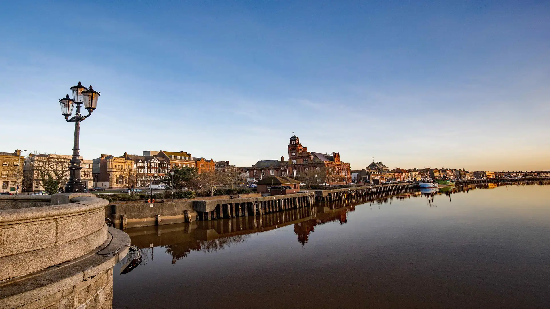 View of Great Yarmouth’s historic quayside at sunset, with period buildings reflecting in the calm river and a traditional lamppost in the foreground.