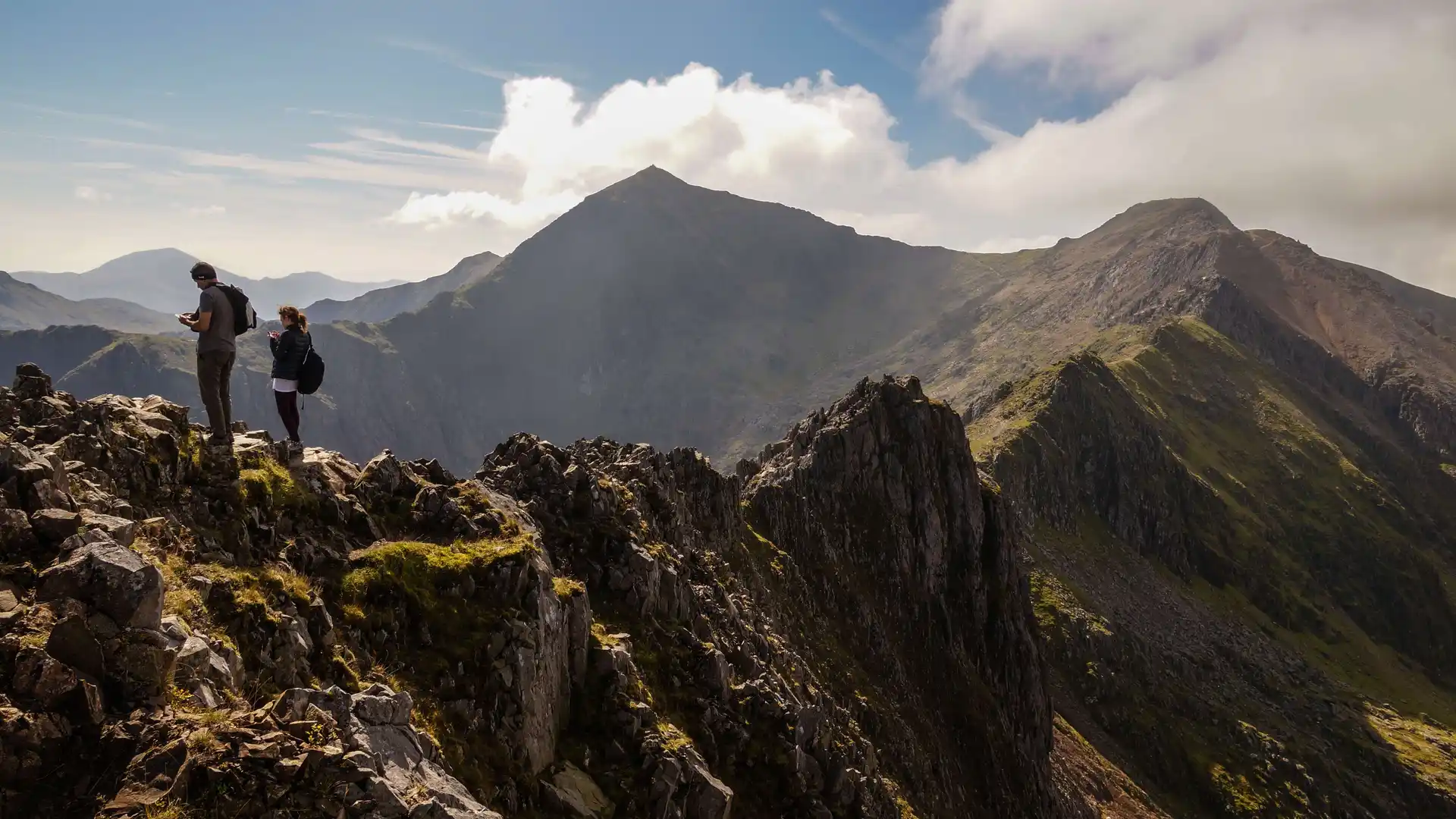 Two hikers standing on a rocky ridge with the summit of Snowdon (Yr Wyddfa) in the distance, surrounded by dramatic mountain peaks under a partly cloudy sky.