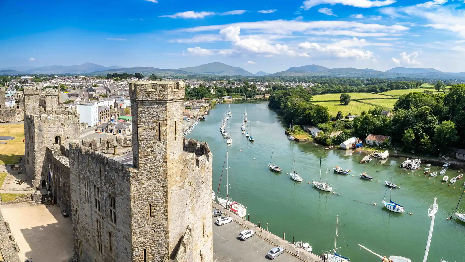 View of Caernarfon Castle in Gwynedd, North Wales, overlooking the River Seiont with sailing boats and distant mountain views under a bright blue sky.