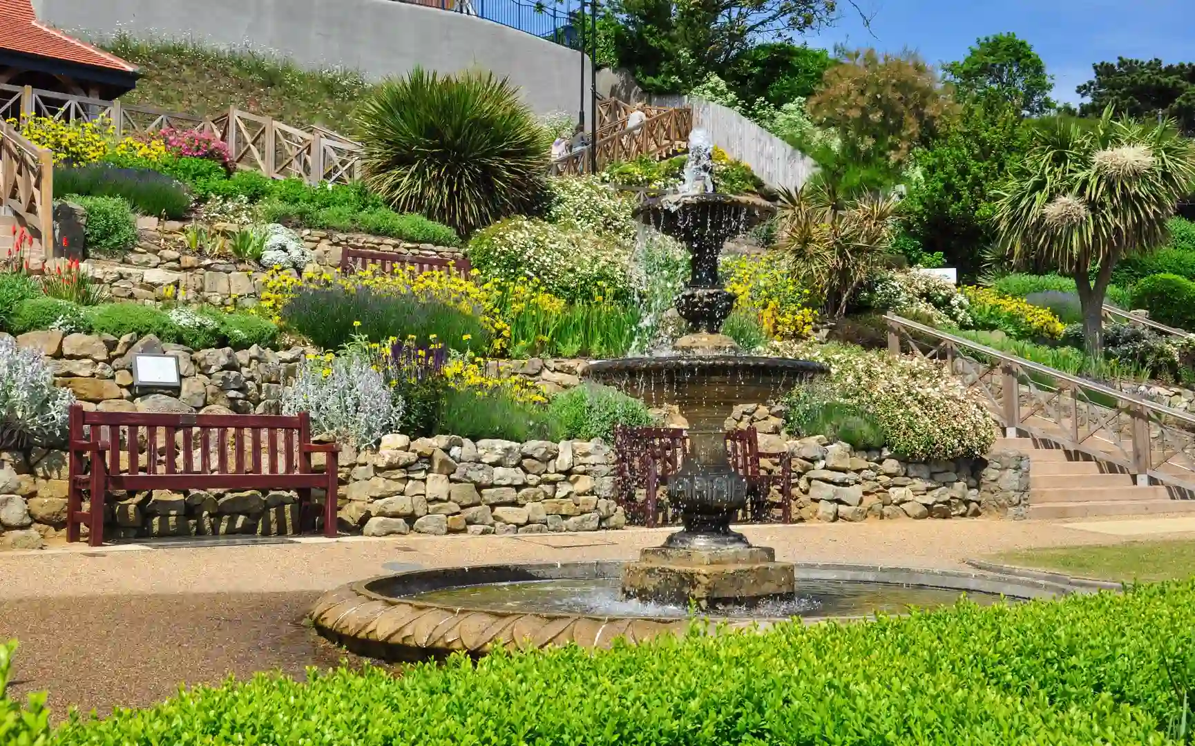 Tiered ornamental fountain surrounded by stone walls, benches, and vibrant flower beds in a landscaped garden on a sunny day.