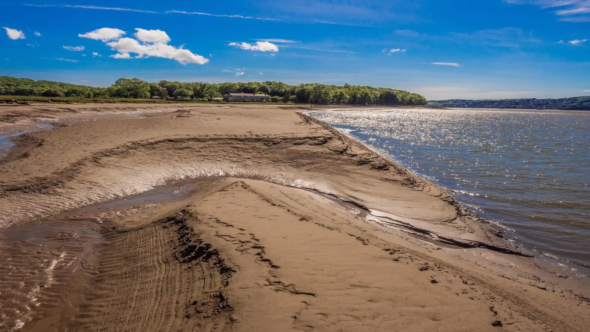 Sandy tidal flats at Arnside with water channels leading into the estuary, backed by green woodland and a bright blue sky.