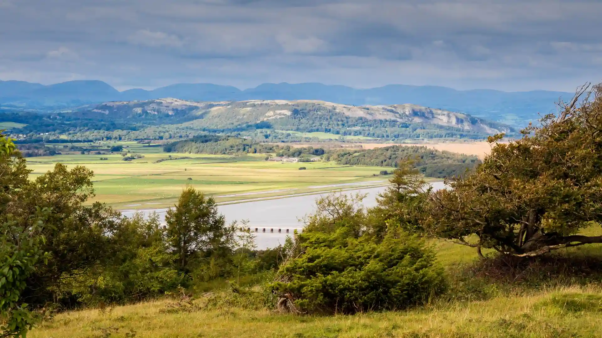 View from Arnside Knott across Morecambe Bay towards wooded hills and limestone ridges, with open fields and estuary below