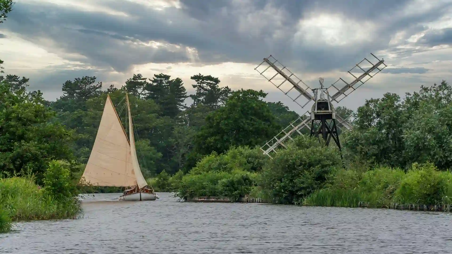 Traditional sailing boat on a river in the Broads with a historic windmill and lush green trees under a cloudy sky.