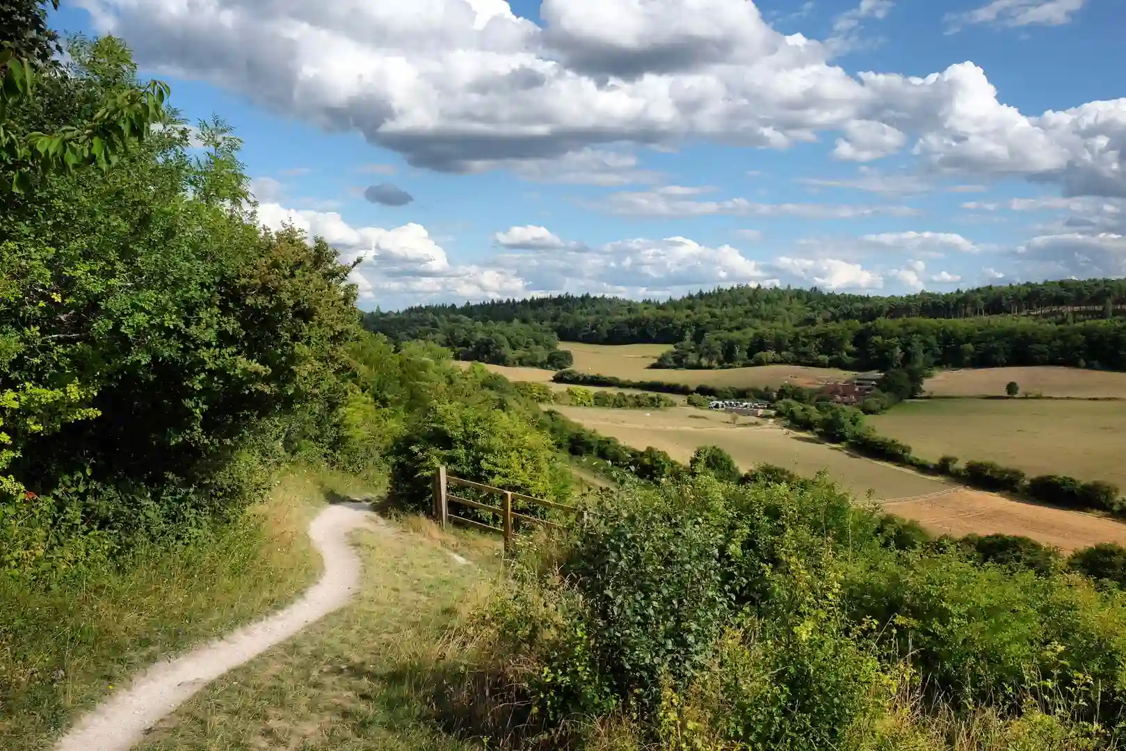 A well worn path leads along side trees and open countryside, with a cloudy blue sky on a summers day.