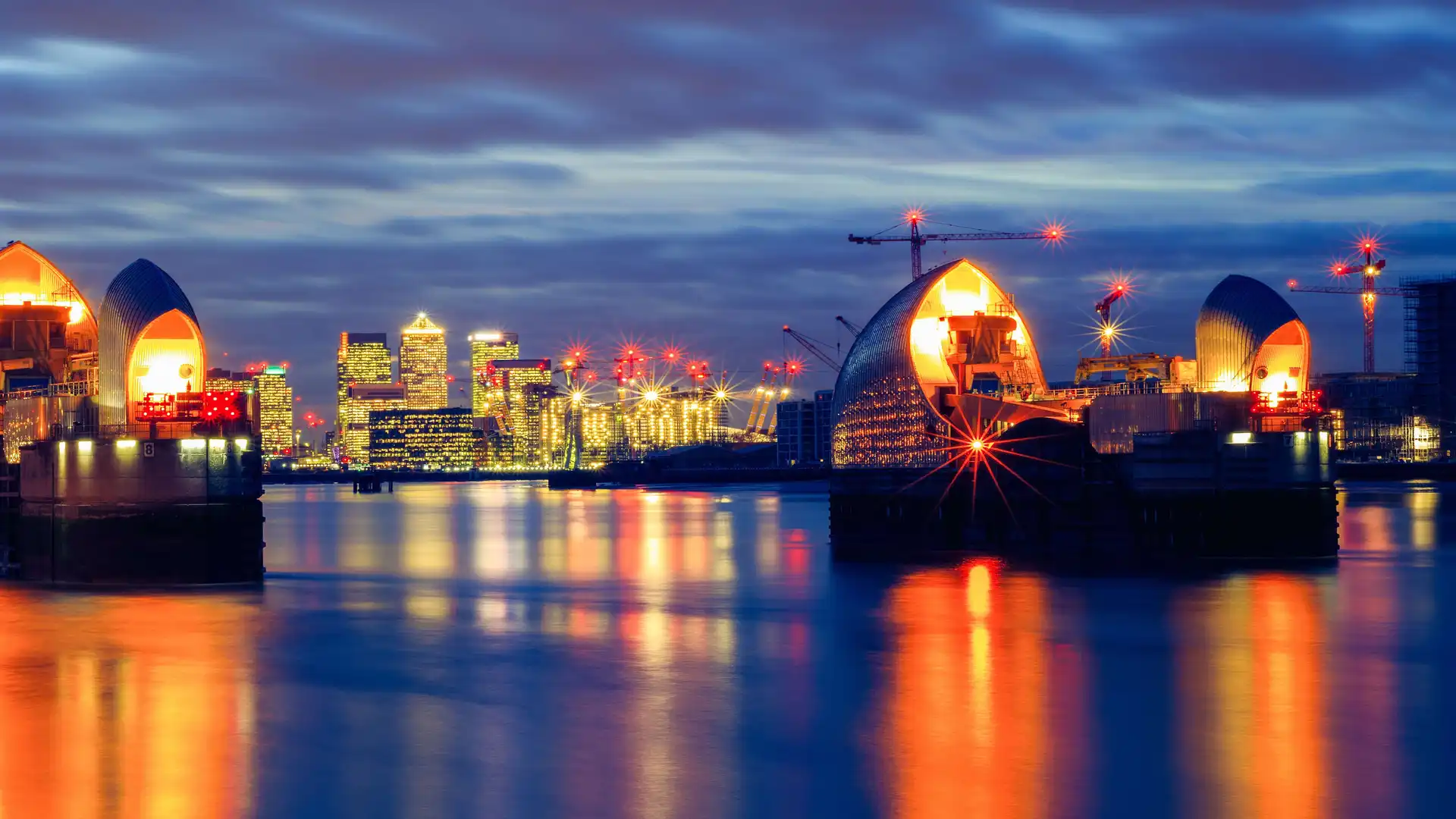 The Thames Barrier illuminated at night with reflections on the River Thames, and the Canary Wharf skyline glowing in the background, London.