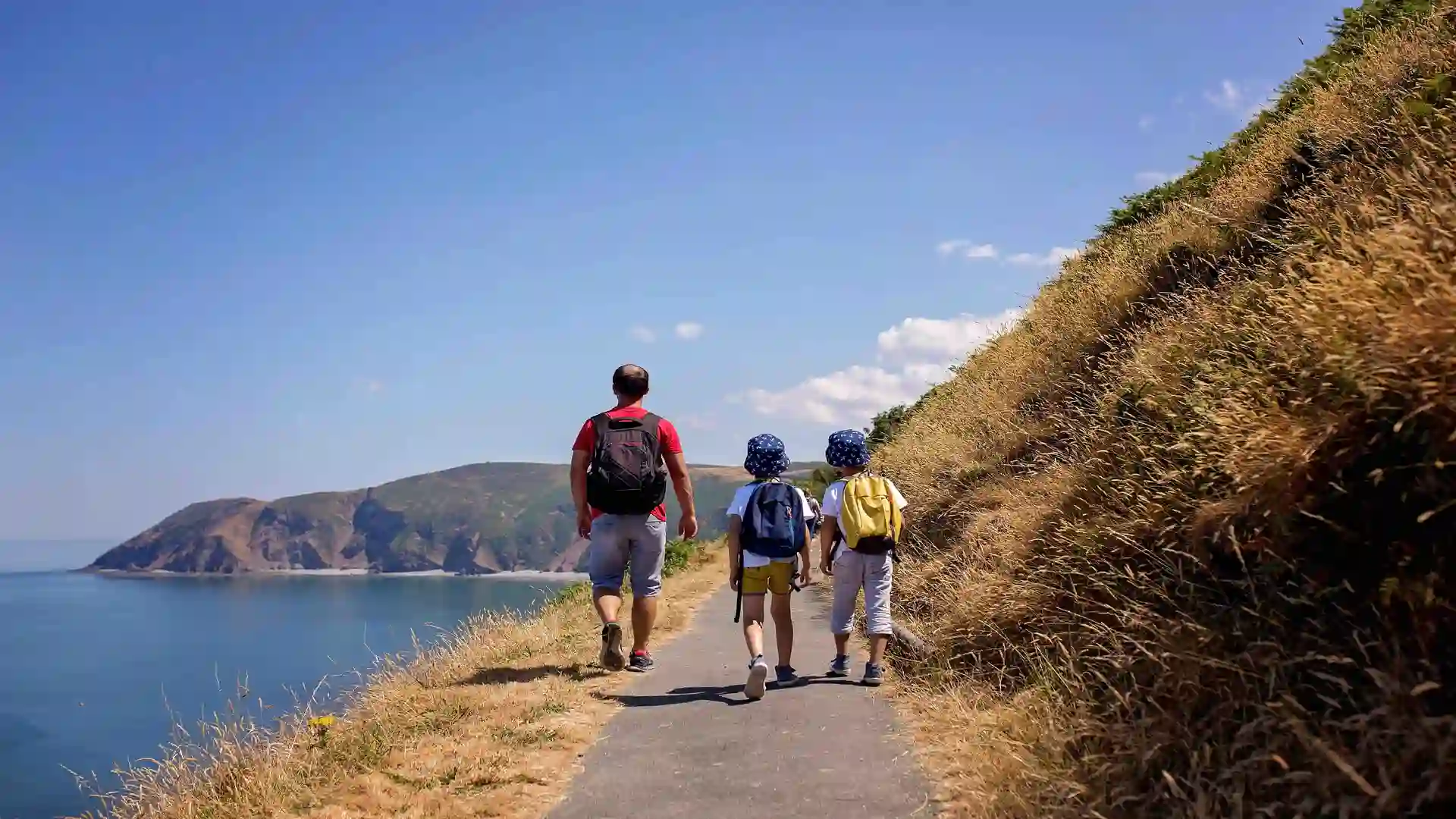 Father and children hiking on the South West Coast Path in Devon, England, with sea cliffs and coastal views in the background.