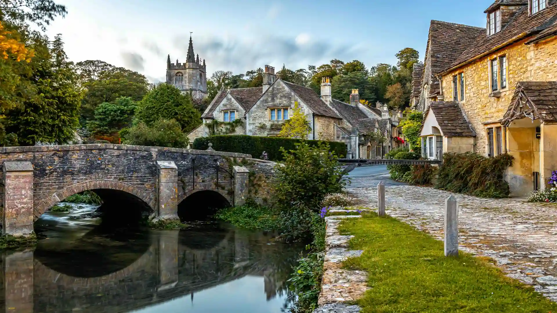 Quaint stone cottages and arched bridge over a stream in Castle Combe village, Wiltshire, England, with the church tower rising in the background.