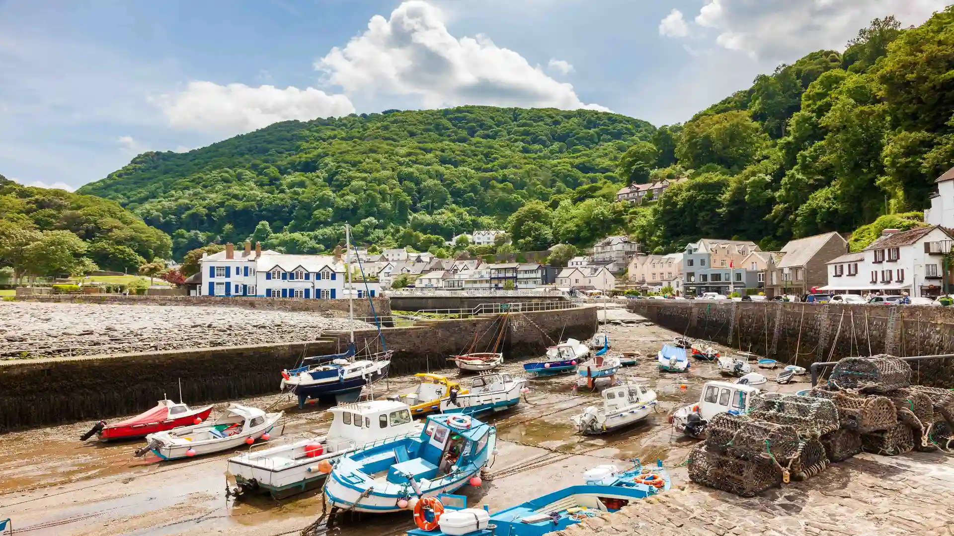 Fishing boats at low tide in Lynmouth harbour, with stone quays, seaside cottages, and tree-covered hills in Exmoor National Park, England.