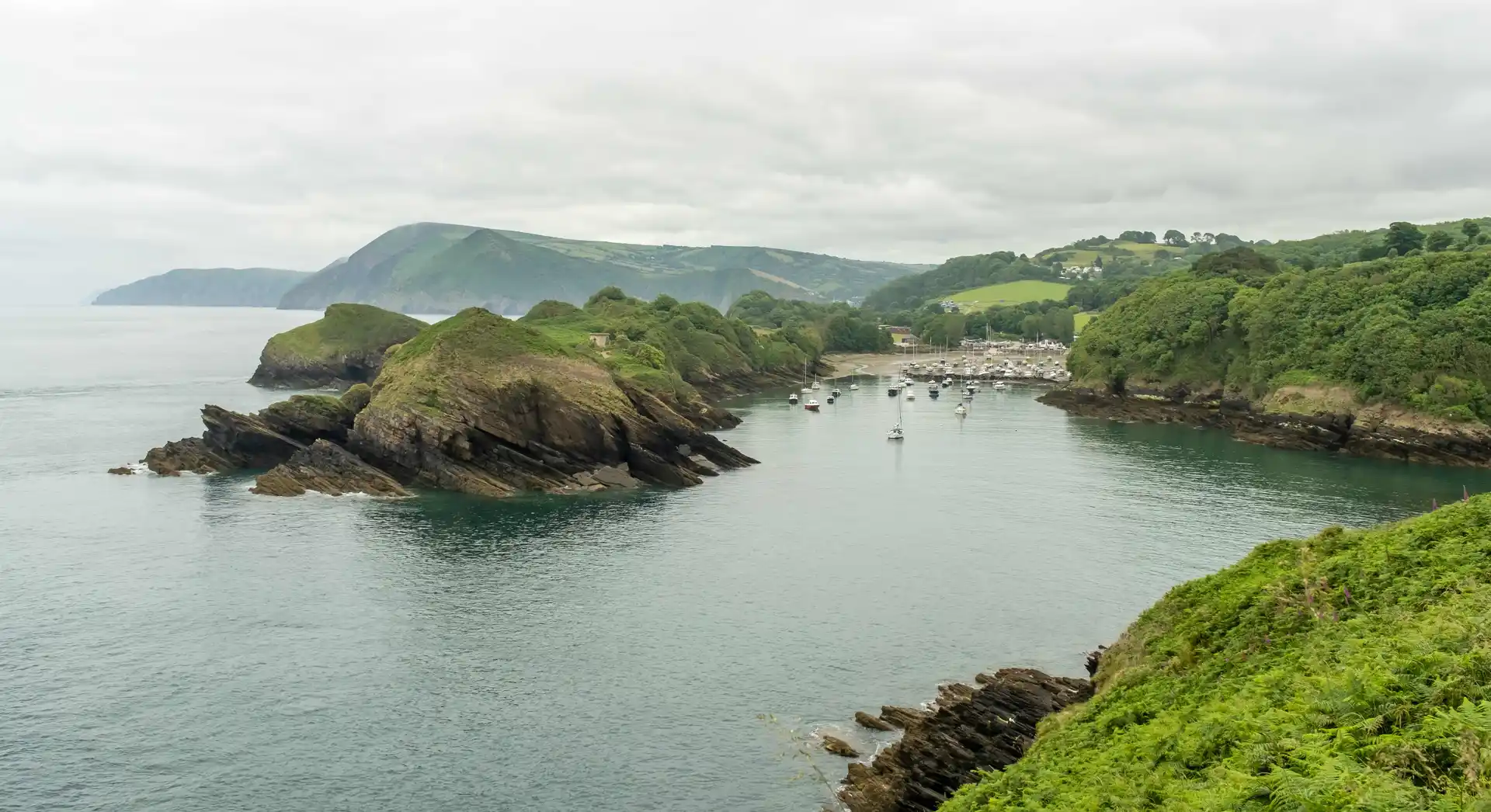 View of Watermouth Harbour and cove from the South West Coast Path in South Devon National Landscape, with turquoise water, moored boats, and wooded headlands.