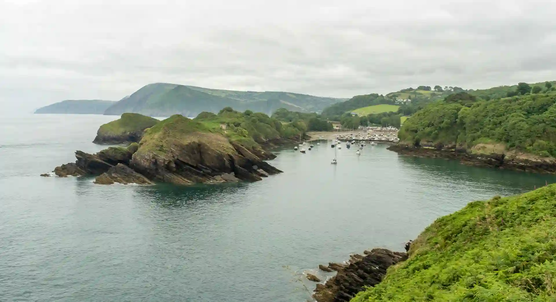 View of Watermouth Harbour and cove from the South West Coast Path in South Devon National Landscape, with turquoise water, moored boats, and wooded headlands.