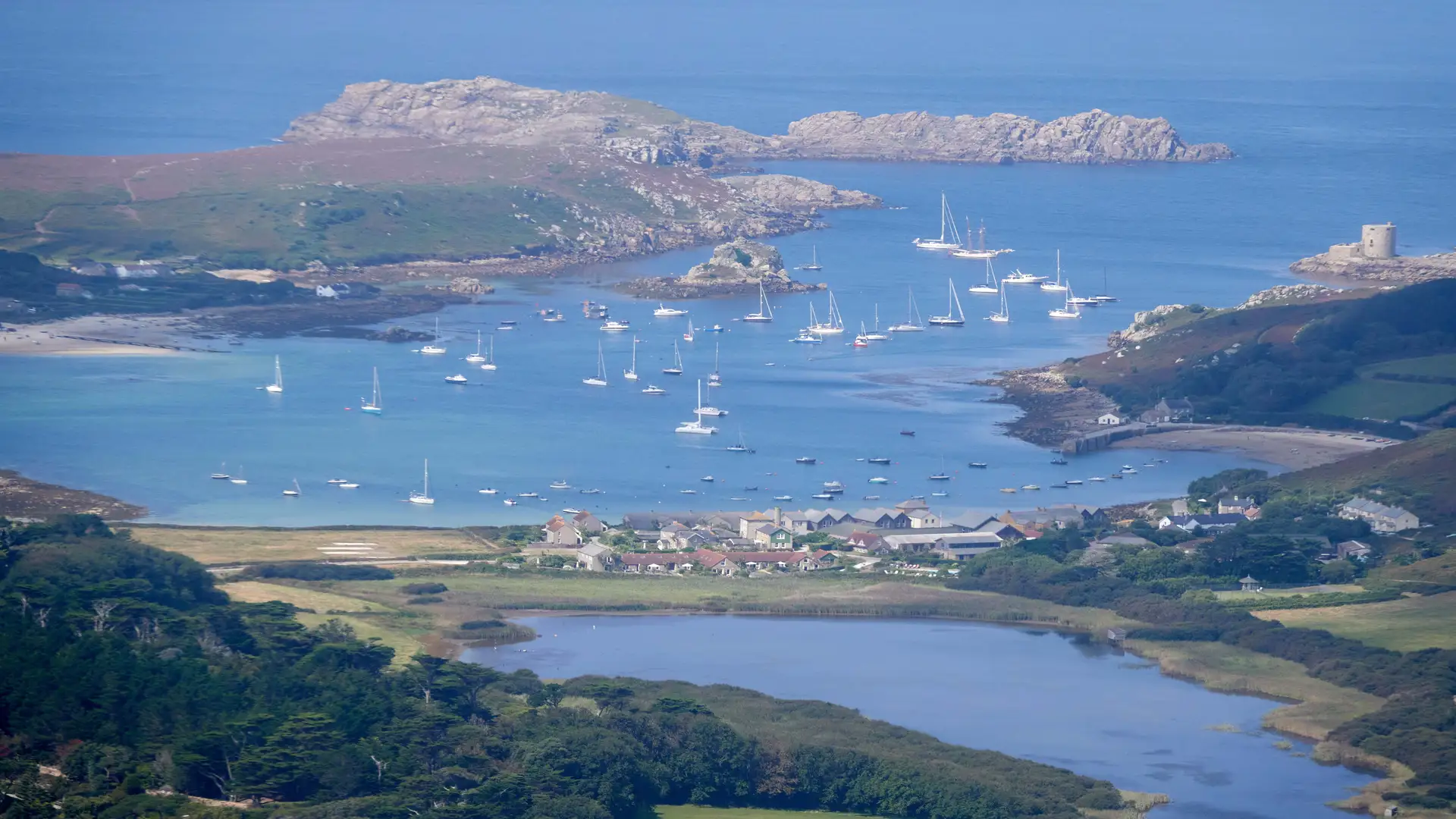 View over the Isles of Scilly National Landscape with boats anchored in a turquoise bay, granite headlands, and small island settlements.