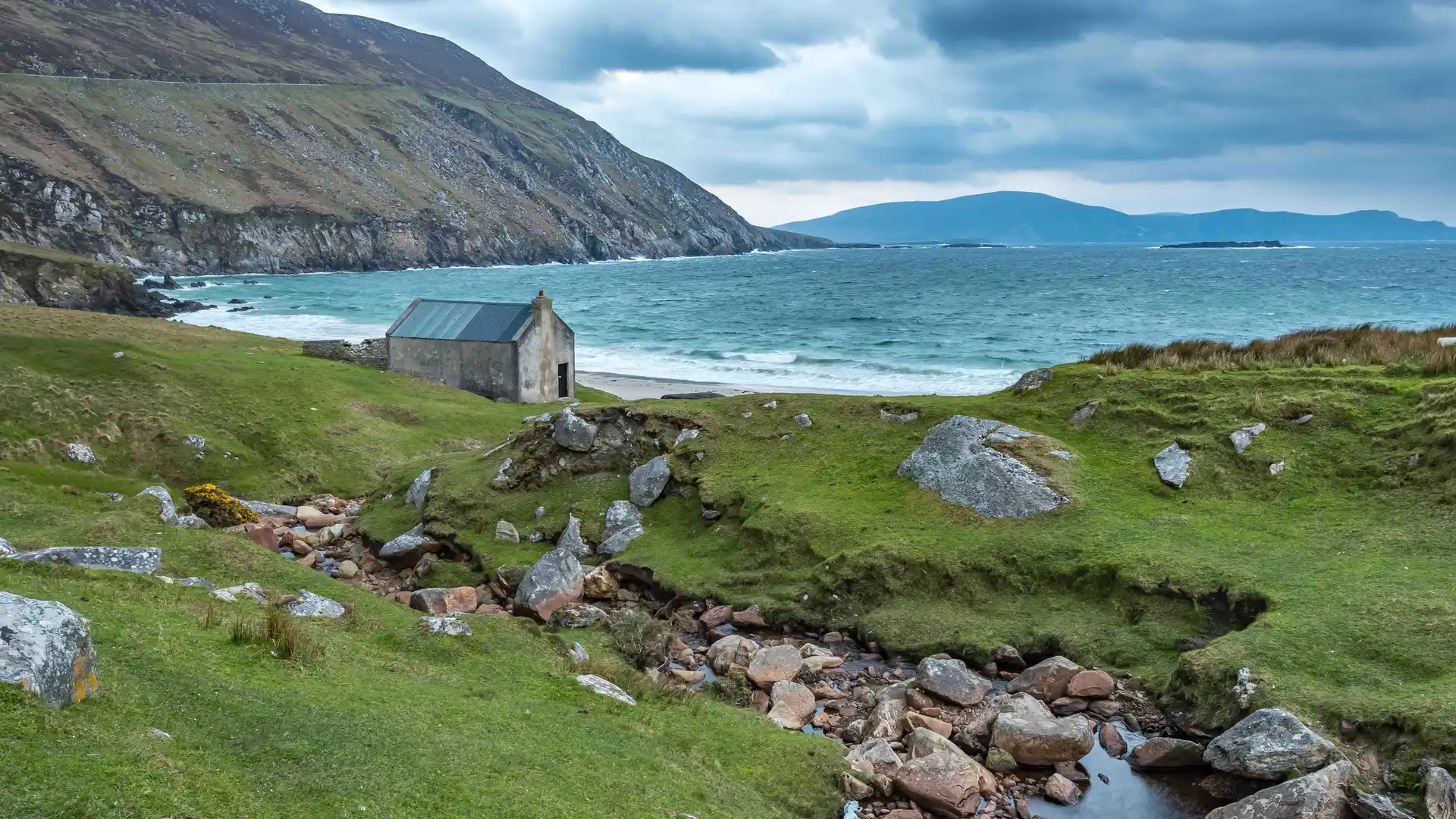 Remote stone cottage at Keem Bay on Achill Island, County Mayo, with green hills, rocky stream, and Atlantic waves in the background.
