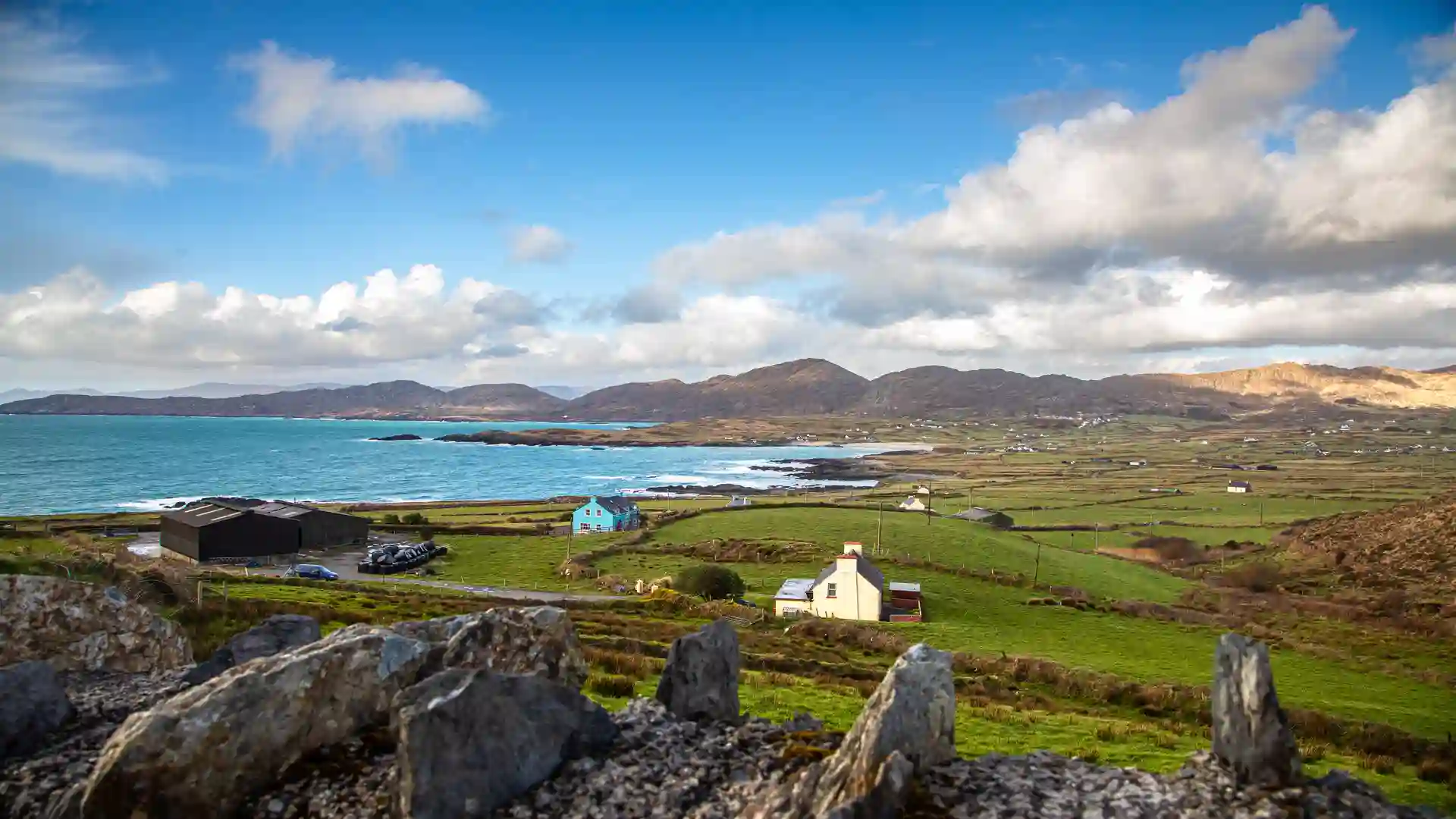 Scenic view over the Beara Peninsula in County Cork, Ireland, with colorful cottages, green fields, rugged mountains, and the Atlantic Ocean under a bright sky.