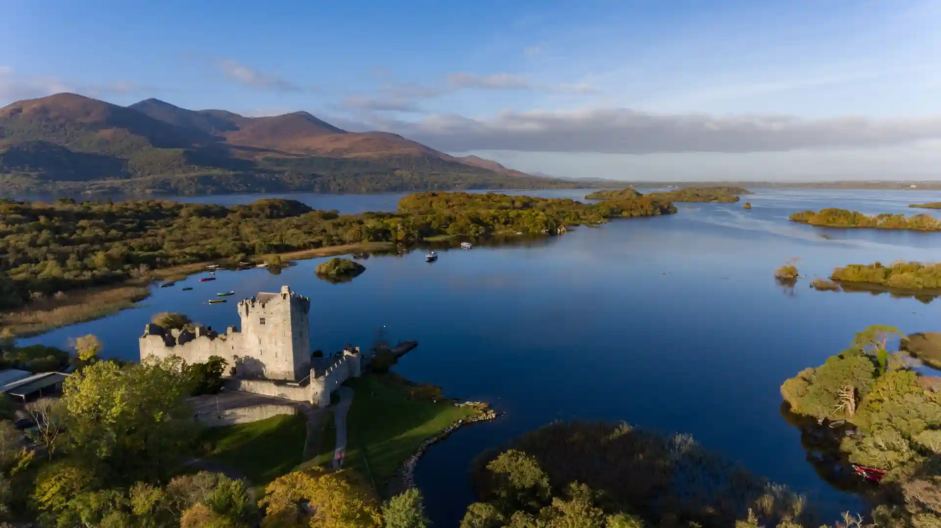 Aerial view of Ross Castle on the shore of Lough Leane in Killarney National Park, County Kerry, with forested islands and the MacGillycuddy’s Reeks mountains in the background.