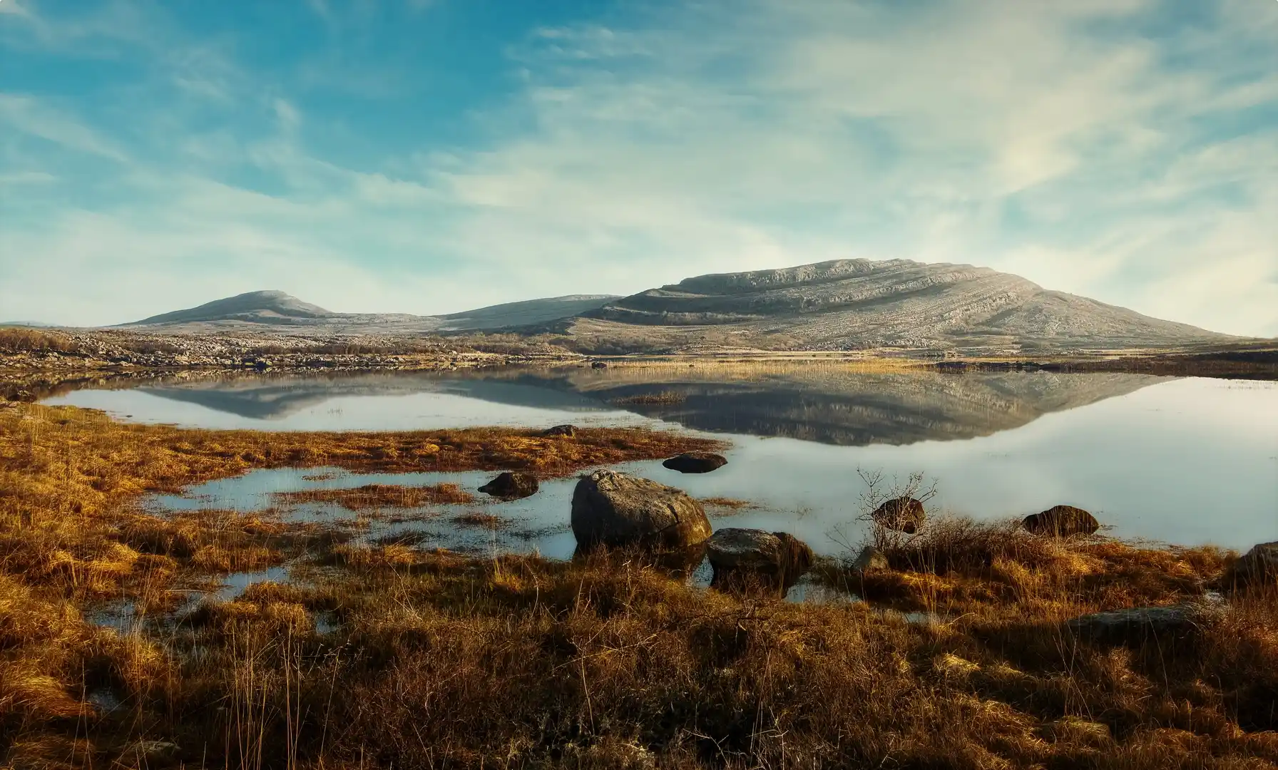 Limestone hills reflected in a still lake at The Burren National Park, County Clare, Ireland, with golden grasses in the foreground under a blue sky.