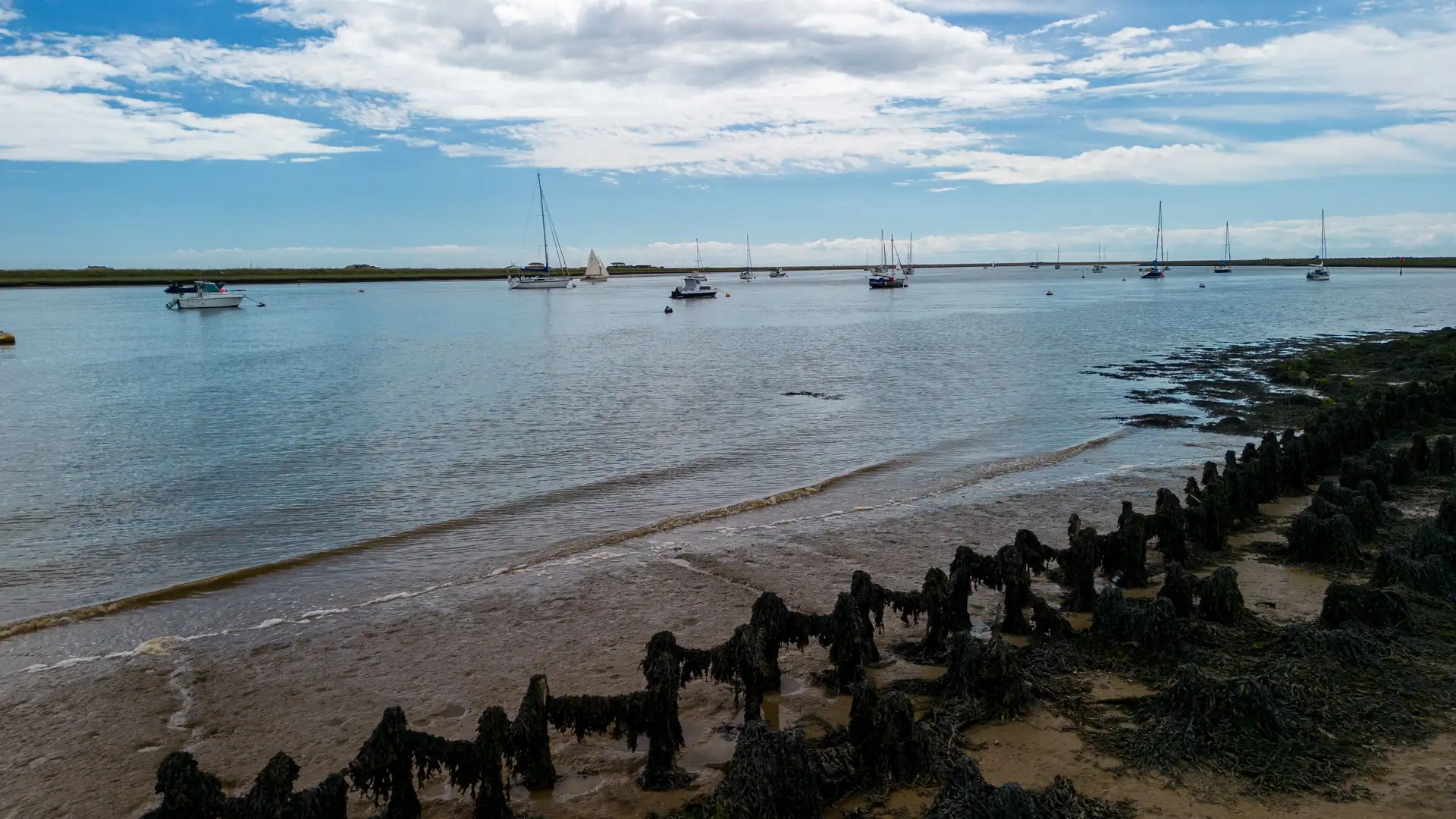 Boats moored on the calm waters of the River Ore at Orford, Suffolk, with seaweed-covered wooden posts in the foreground and blue skies above.