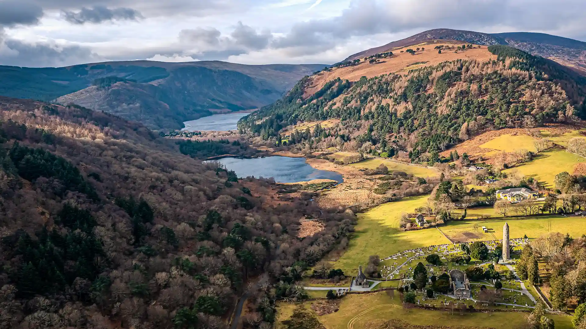 Aerial view of Glendalough Valley in County Wicklow, Ireland, showing the twin lakes surrounded by forested hills and the early medieval monastic site with its round tower — a highlight along the Wicklow Way walking trail.