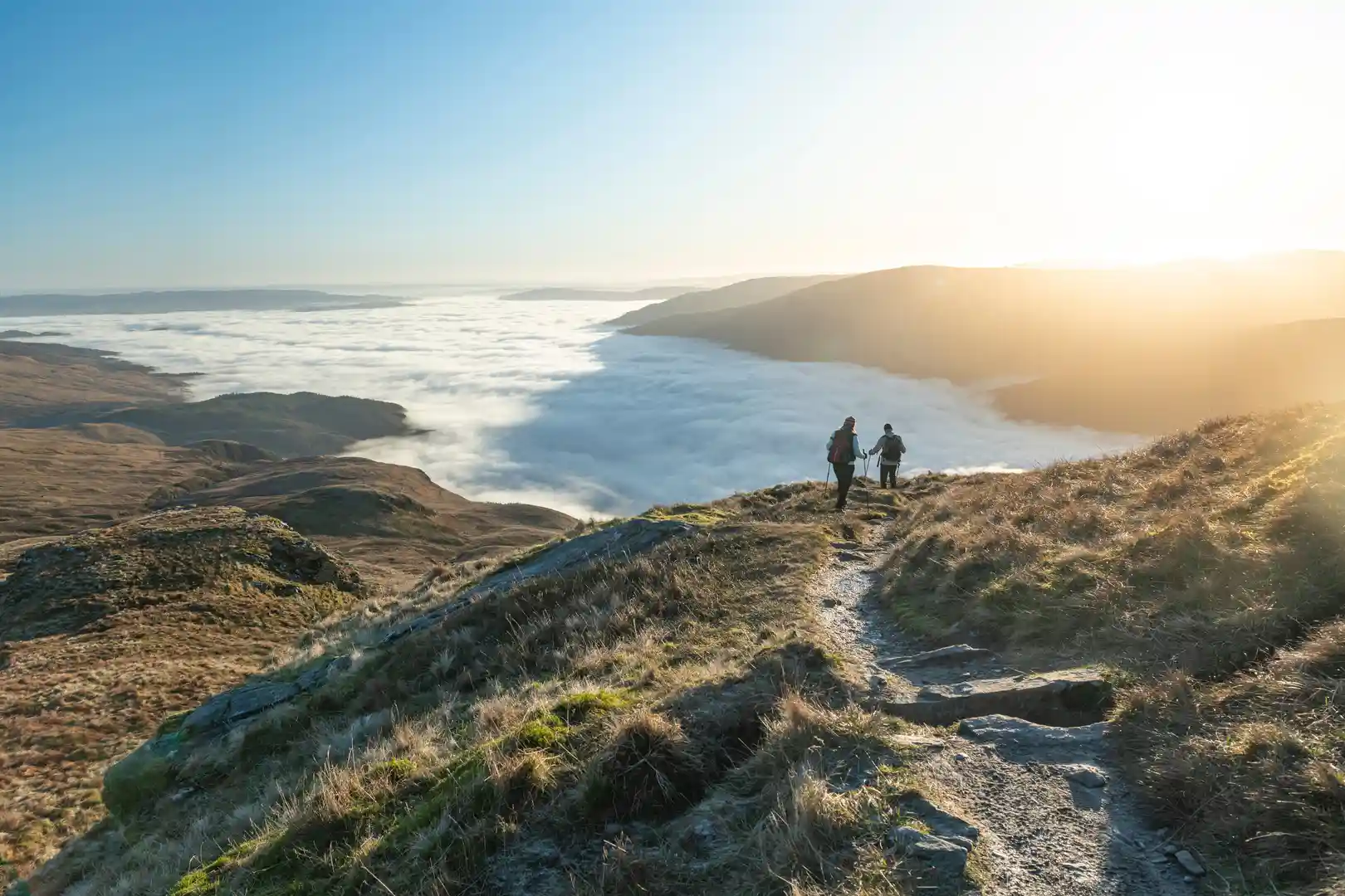 Two hikers walk along a mountain trail above a sea of clouds in Loch Lomond & The Trossachs National Park, Scotland, with golden morning light over the surrounding hills.
