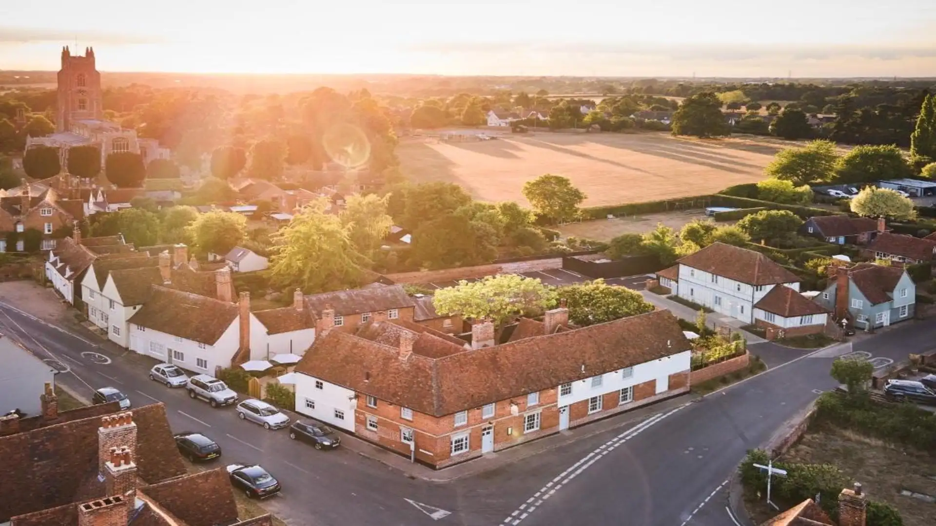 Aerial view of Stoke-by-Nayland village in Suffolk at sunset, showing red-brick cottages, the parish church, and surrounding countryside within the Dedham Vale National Landscape.