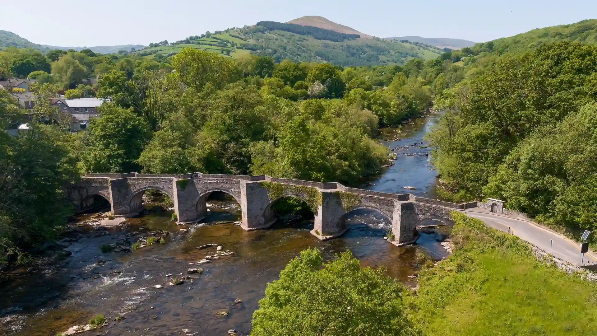 Stone bridge crossing the River Usk at Crickhowell in Powys, surrounded by green hills and trees with the Brecon Beacons in the distance.