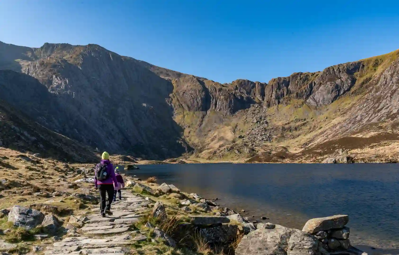 Walkers on a mountain trail beside Llyn Idwal in Eryri (Snowdonia) National Park, surrounded by rugged peaks and clear blue skies in North Wales.