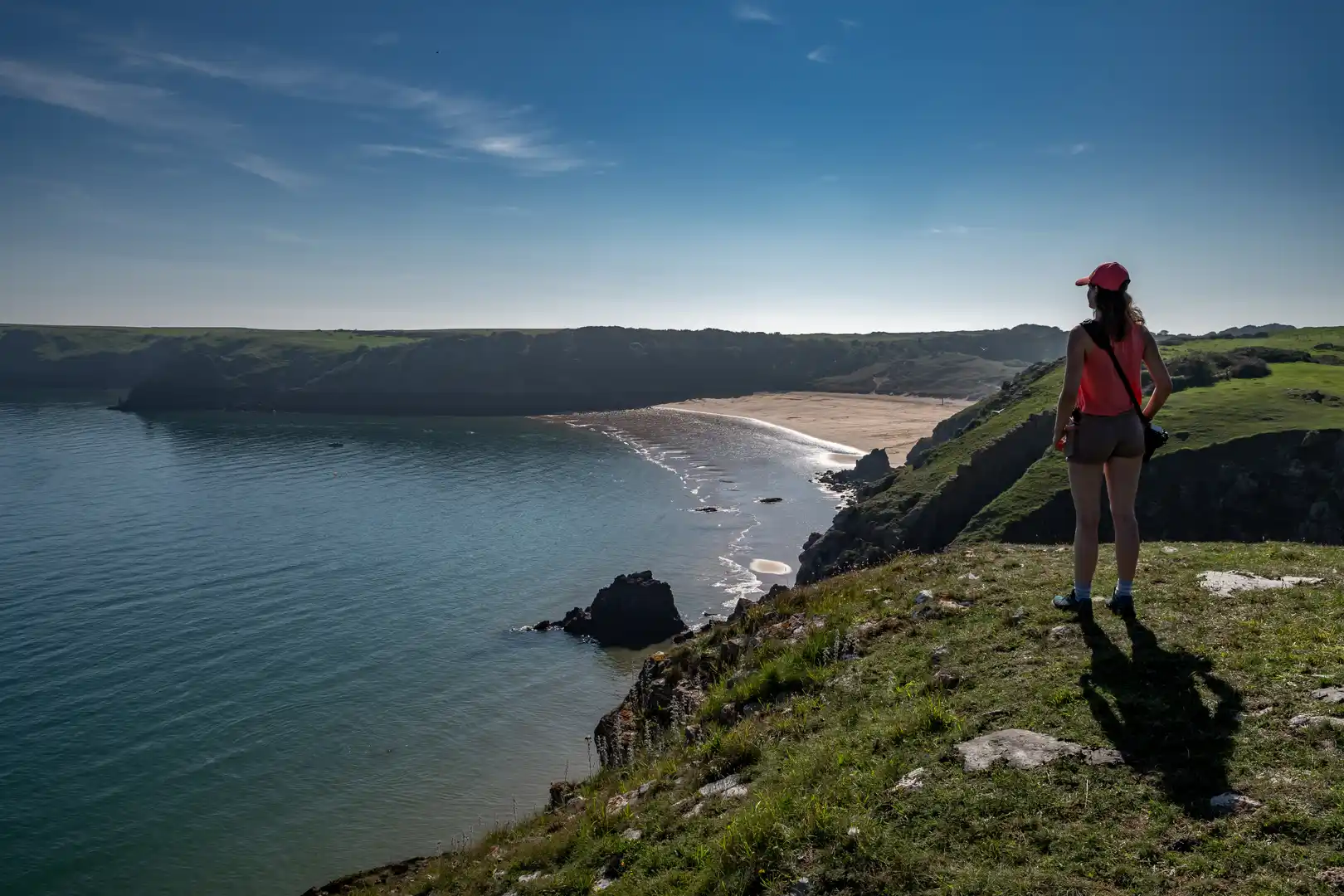 A walker stands on a grassy cliff overlooking a sweeping sandy bay along the Pembrokeshire Coast Path in Wales, with calm blue sea and rugged headlands under a clear sky.