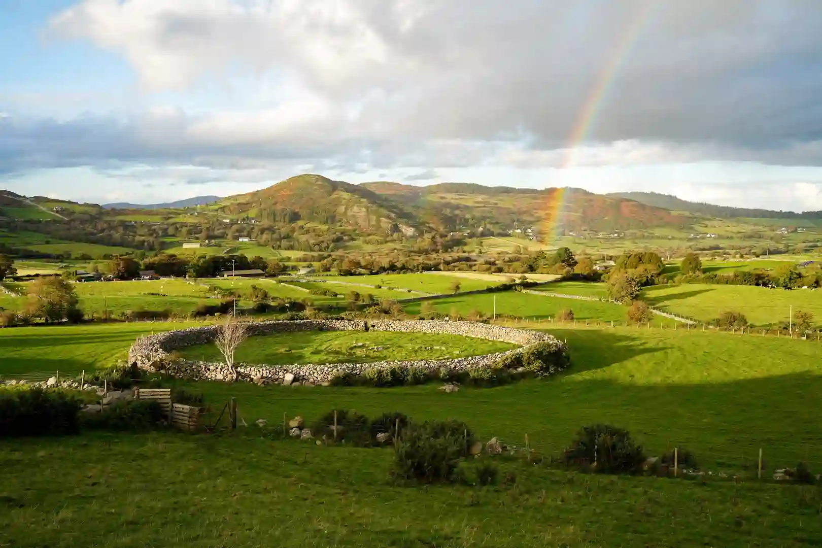 View over the Ring of Gullion in County Armagh, Northern Ireland, showing green fields, stone walls, and a rainbow over rolling hills — a peaceful walking landscape.