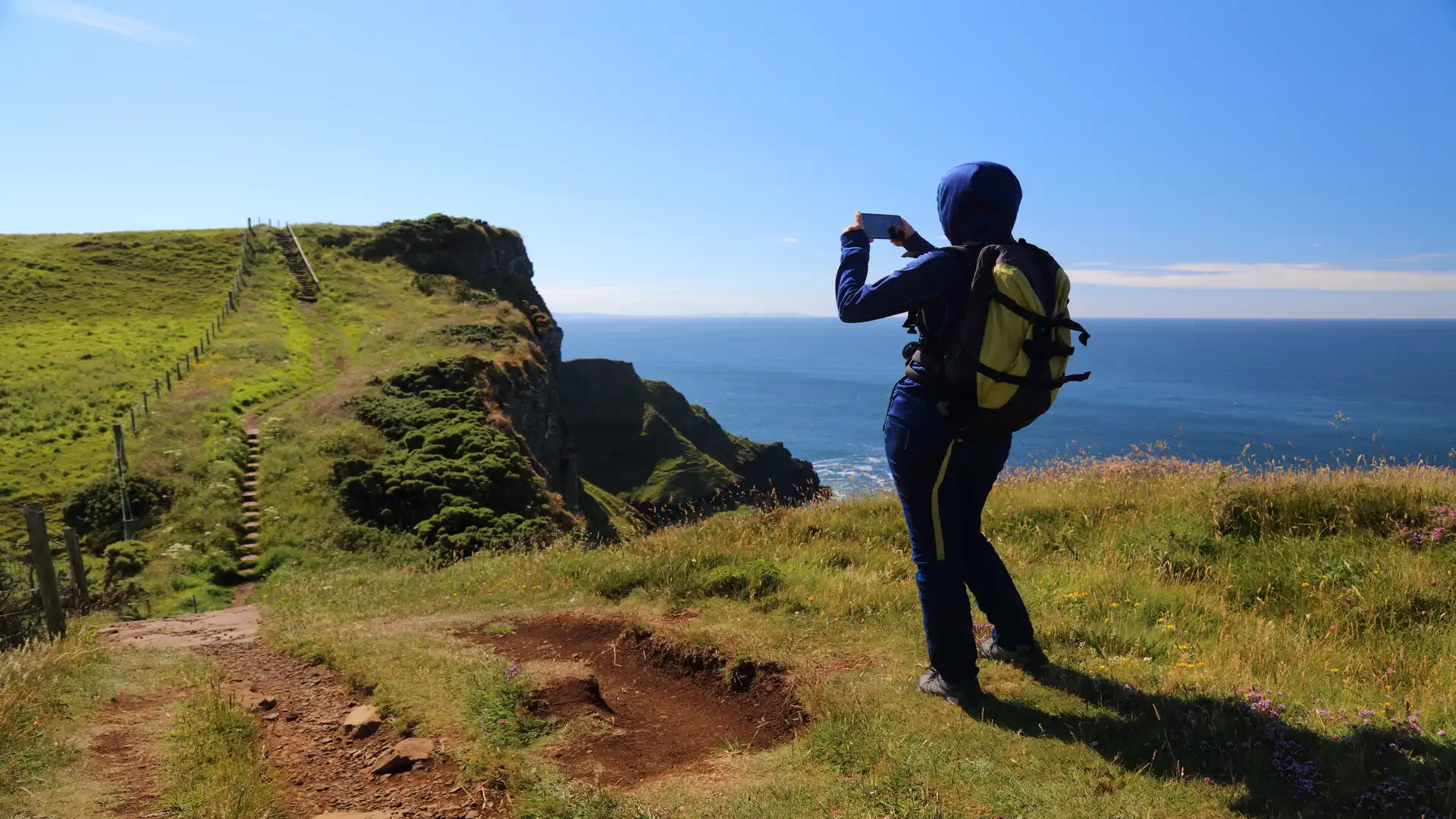 Hiker photographing the clifftop path along the Causeway Coast Way in Northern Ireland, overlooking the Atlantic Ocean on a sunny day.