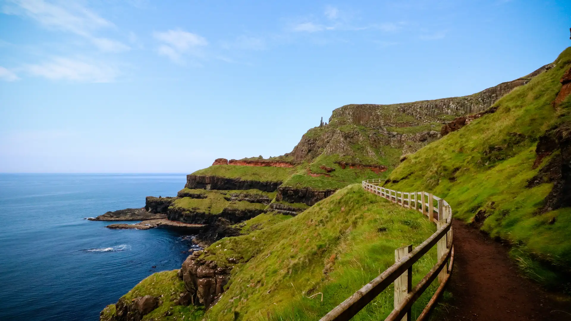 Clifftop walking path overlooking the Atlantic Ocean along the Causeway Coast in Northern Ireland, with green hills and layered basalt cliffs under a bright blue sky.