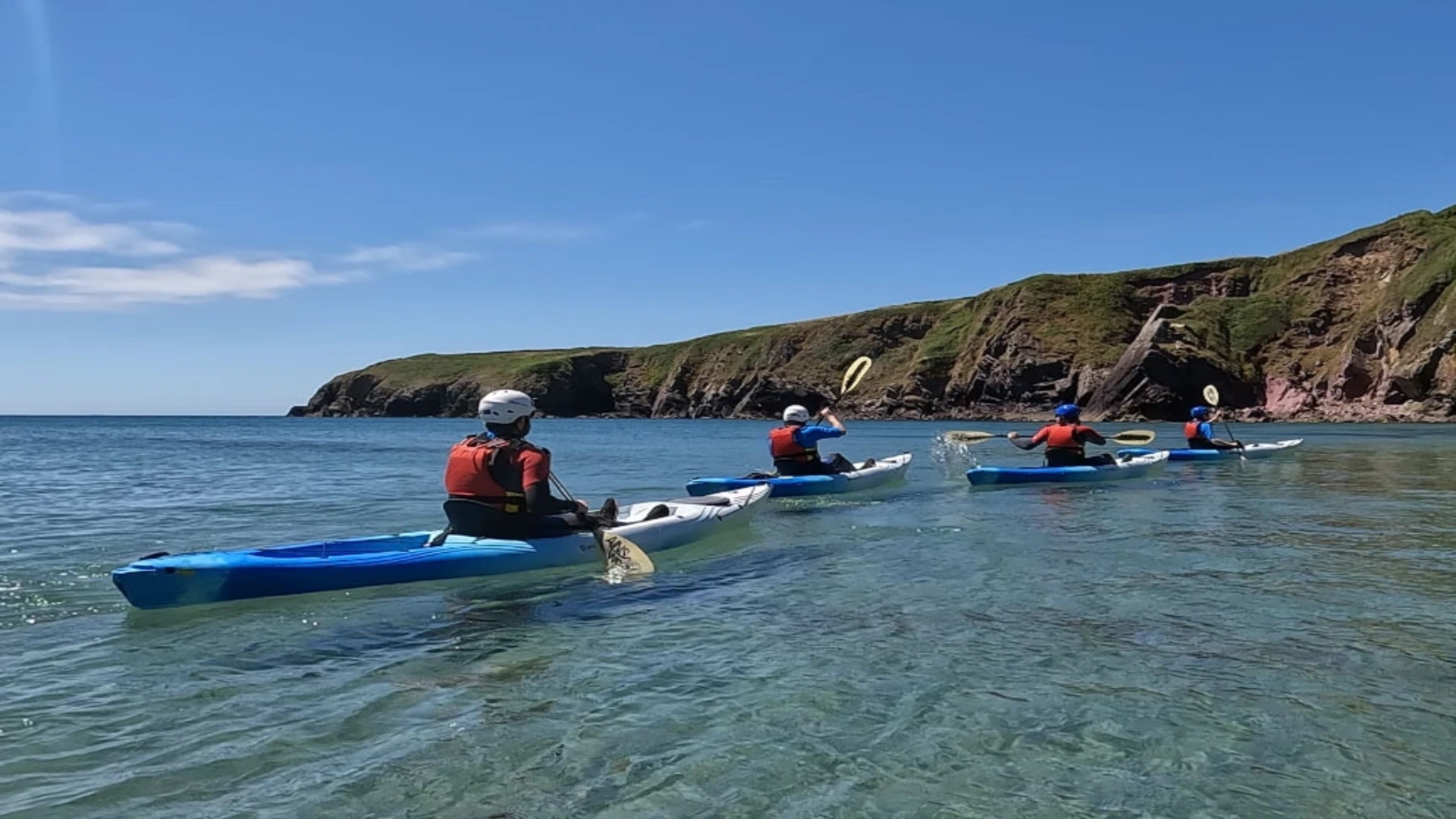 Group of people sea kayaking along a clear, calm bay beneath rugged coastal cliffs on a sunny day in Pembrokeshire, Wales.