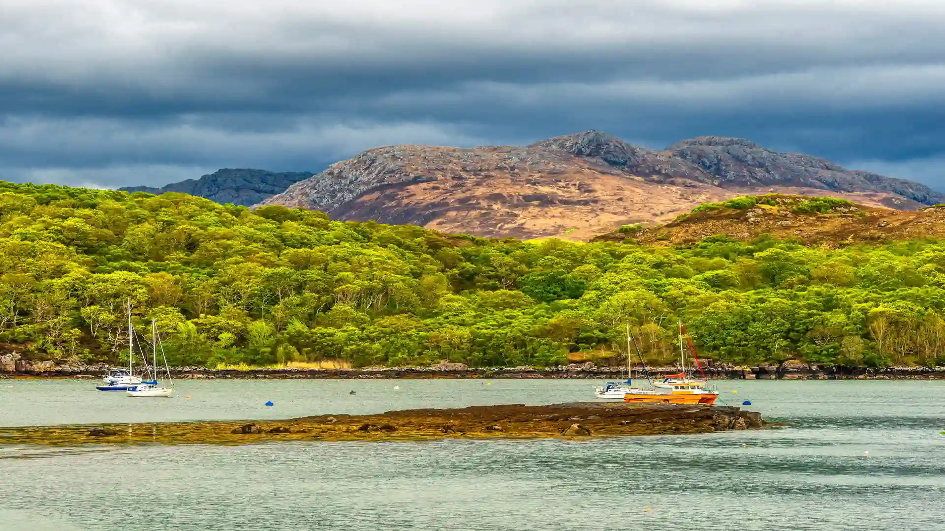 View across Gairloch towards the rugged mountains of Wester Ross in the Scottish Highlands, with yachts anchored in calm waters beneath dramatic skies.