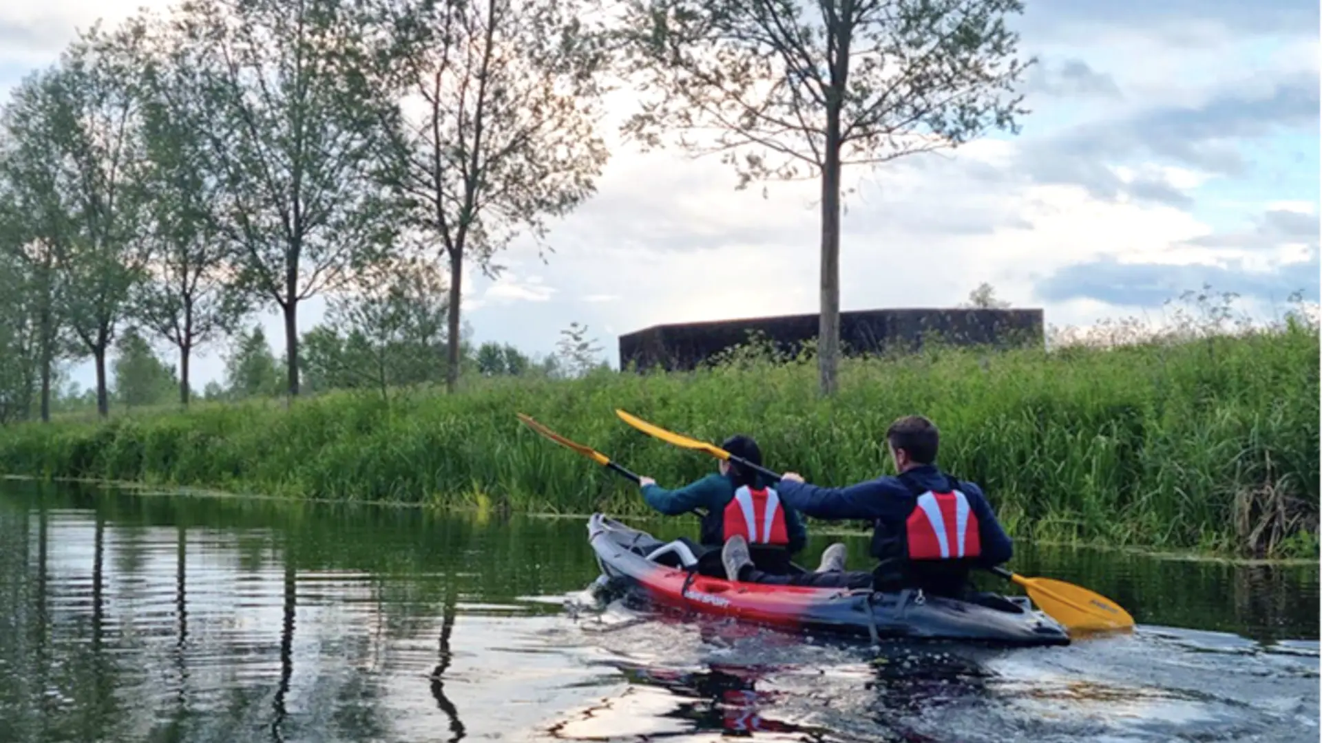 Two people kayaking on the River Stour surrounded by tall grasses and trees in the Dedham Vale National Landscape, with calm reflections on the water at sunset.